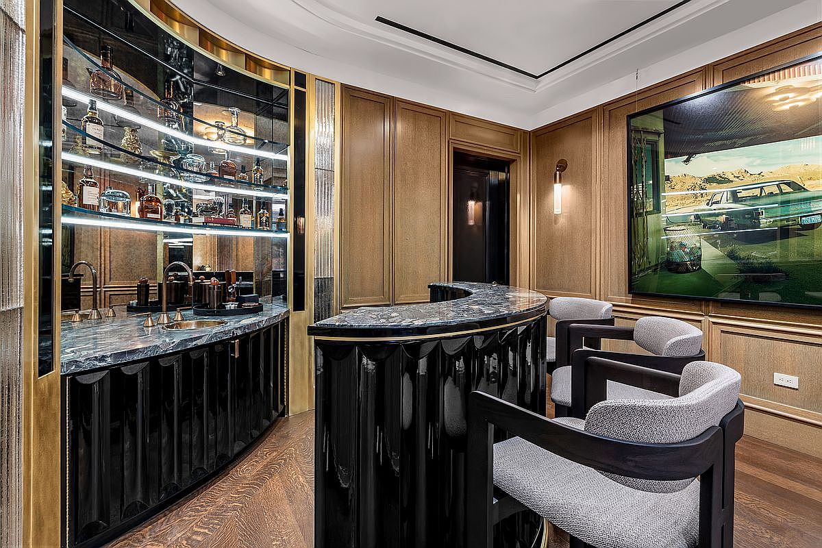 This sophisticated home bar area features a curved, high-gloss black bar with a dark stone countertop and matching bar stools. Behind the bar, a backlit glass shelving unit displays an array of spirits, framed by gold-toned metal accents and wood-paneled walls. A large, framed photograph of a vintage car adds a touch of character to the room, which is finished with rich wood flooring and elegant wall sconces.
