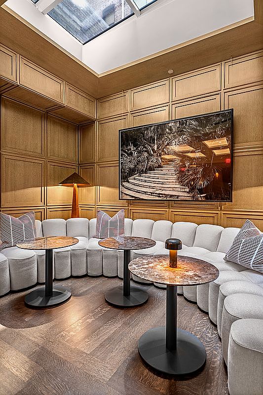 This sophisticated lounge area features a curved, light-colored sectional sofa arranged around three circular tables with dark stone tops. The walls are adorned with wood paneling, and a large, framed black-and-white photograph hangs above the seating area, while a skylight above provides natural illumination. The space is finished with dark wood flooring, creating a warm and upscale atmosphere suitable for a high-end residential or commercial lobby.