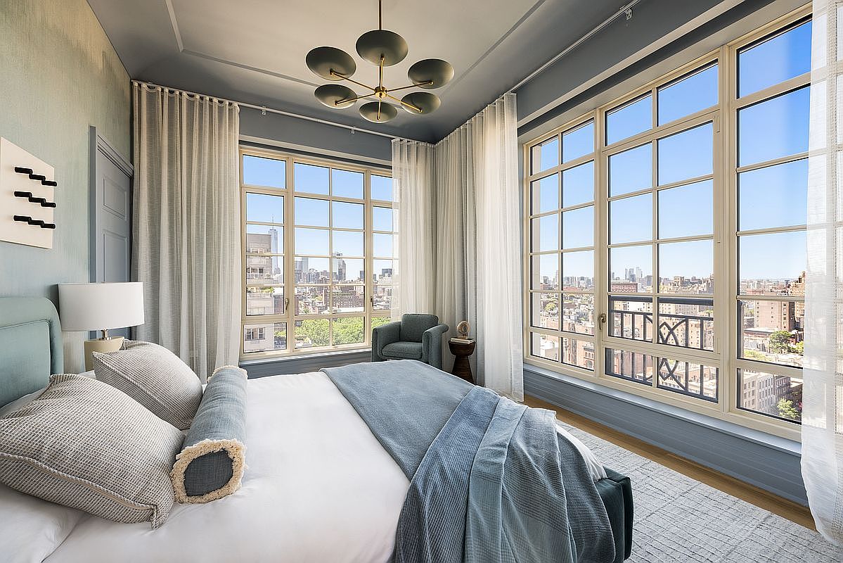 This elegant primary bedroom features a sophisticated blue and white color palette, centered around a plush bed with layered linens and a textured throw. The room is bathed in natural light from expansive windows that offer sweeping urban views, complemented by sheer curtains and a modern, multi-armed chandelier. A cozy armchair sits in the corner, creating a serene and refined atmosphere suitable for a high-end city residence.