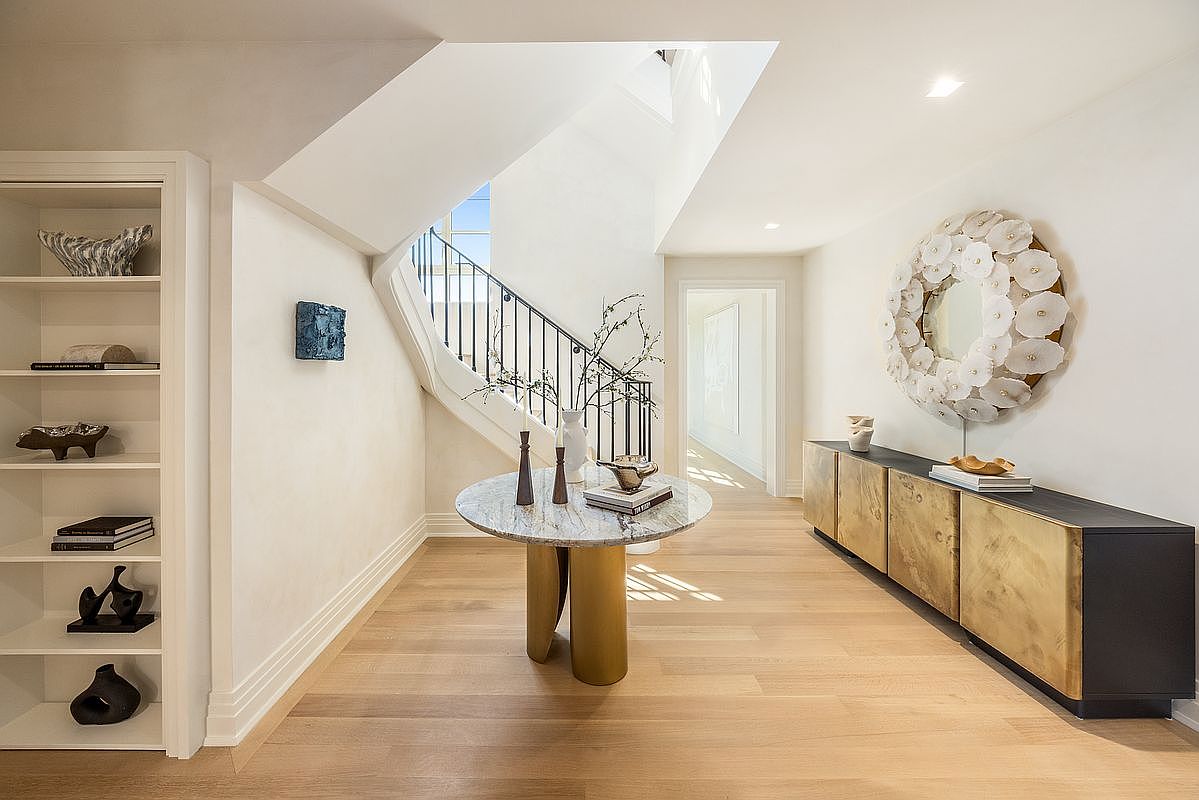 This elegant entryway features a curved staircase with a black metal railing and light wood flooring that extends into a bright hallway. A circular stone-topped pedestal table serves as a central focal point, complemented by a large, textured floral-inspired wall sculpture above a long, metallic-finished console cabinet. The space is bathed in natural light, creating a sophisticated and airy atmosphere that welcomes guests into the home.