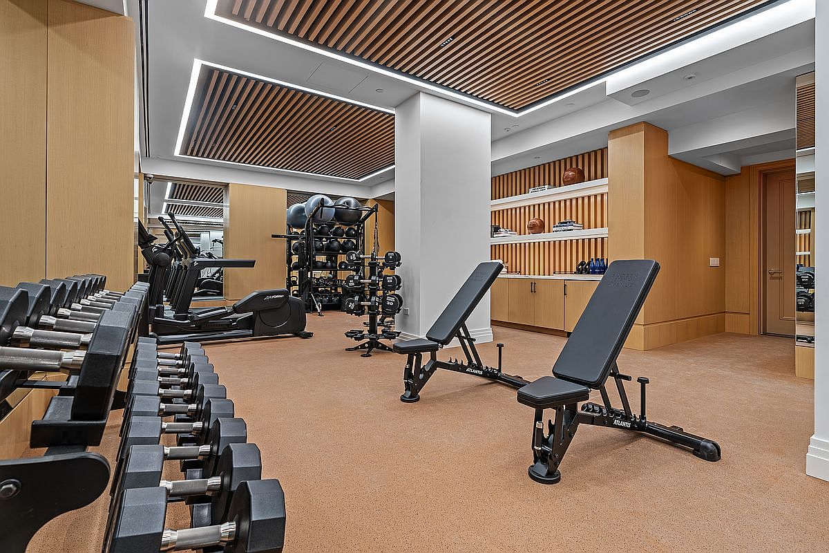 This professional-grade home gym features a clean, modern aesthetic with light wood wall paneling and a slatted wood ceiling design. The space is equipped with a variety of fitness equipment, including a row of dumbbells on a rack, two adjustable weight benches, and cardio machines in the background. The room is well-lit with recessed lighting and integrated linear light strips, creating a bright and inviting atmosphere for exercise.