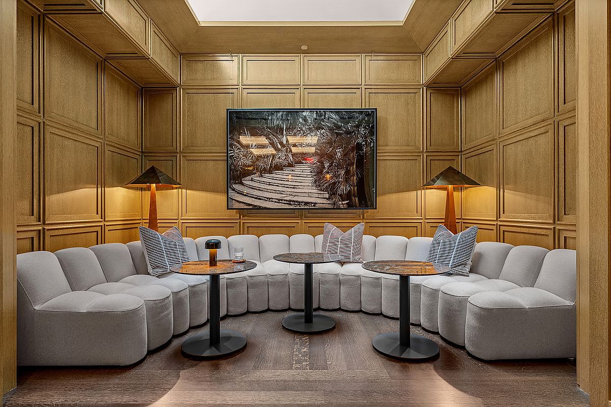 This sophisticated lounge area features a large, curved modular sofa upholstered in light fabric, set against walls paneled in warm-toned wood. Three small, round tables with dark stone tops are arranged in front of the seating, while two metallic lamps provide soft, ambient lighting on either side of a central framed photograph. The space is framed by a recessed ceiling detail, creating an intimate and luxurious atmosphere perfect for social gatherings.