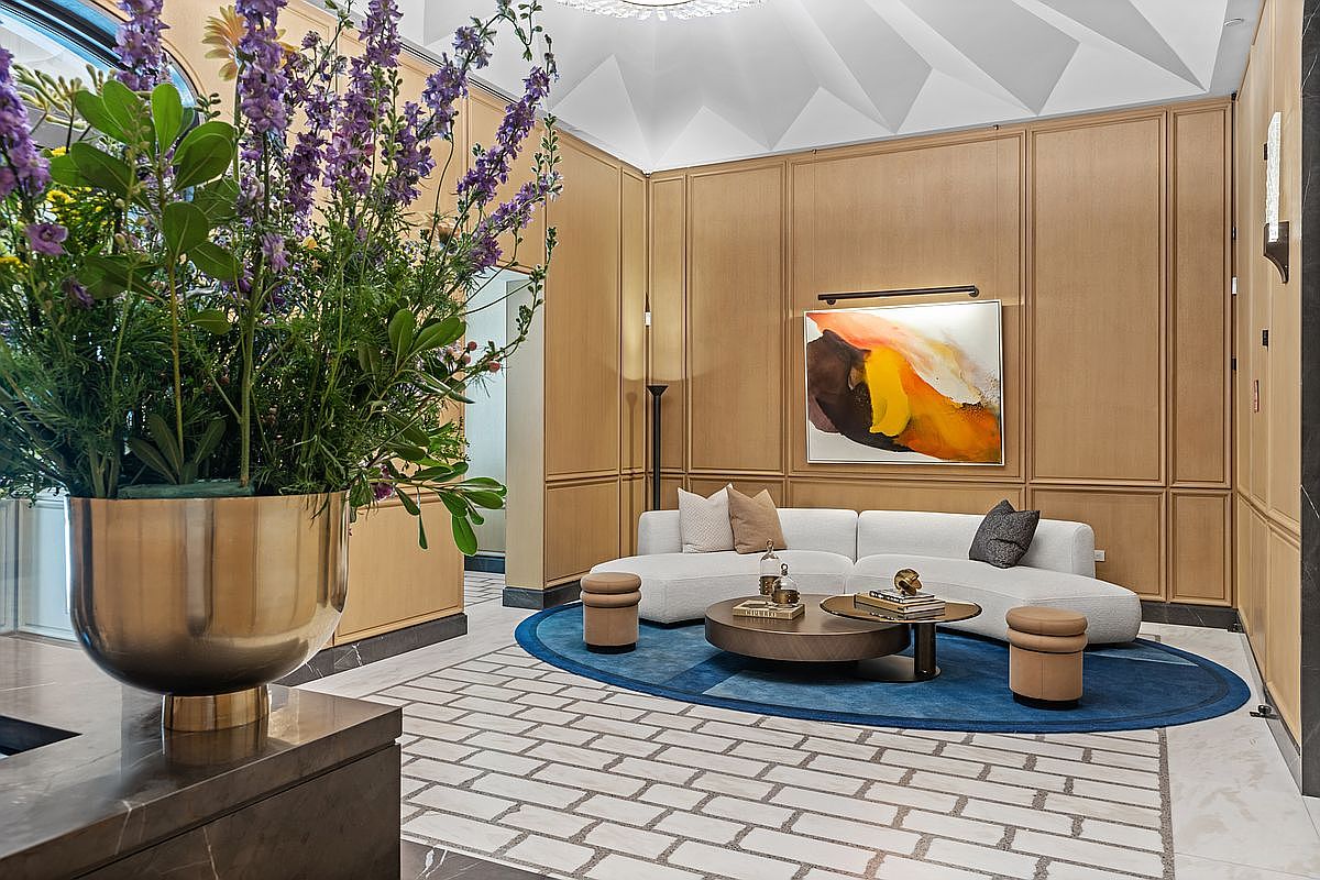 This sophisticated living area features light wood paneling on the walls and a striking, multi-faceted ceiling design. A curved, light-colored sofa sits atop a deep blue oval rug, complemented by a round coffee table and two small ottomans. In the foreground, a large metallic vase filled with purple flowers adds a vibrant touch, while a colorful abstract painting hangs above the sofa, illuminated by a dedicated picture light.