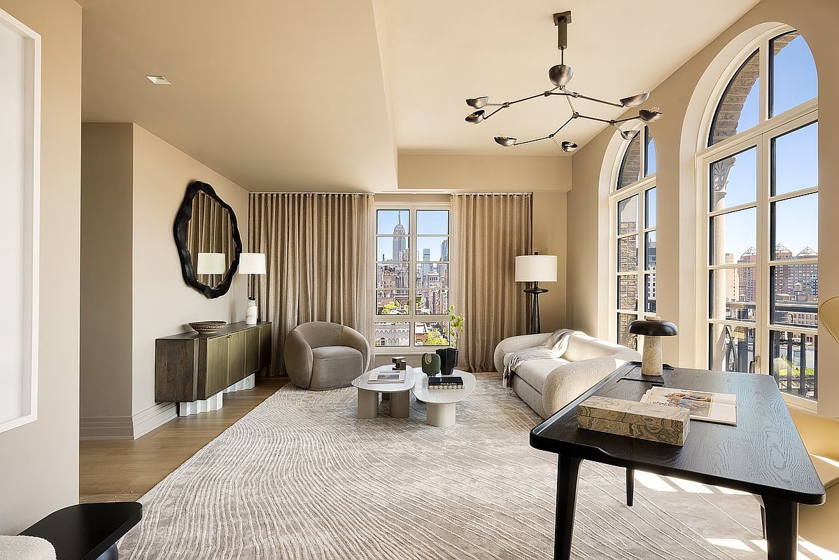 This sophisticated living room features a neutral color palette, a textured area rug, and a mix of modern furniture including a curved sofa and a plush armchair. The space is anchored by a dark wood desk in the foreground and a sleek sideboard against the wall, while large arched windows provide an iconic view of the city skyline. A contemporary multi-arm chandelier hangs from the ceiling, adding a sculptural element to the refined, airy atmosphere.