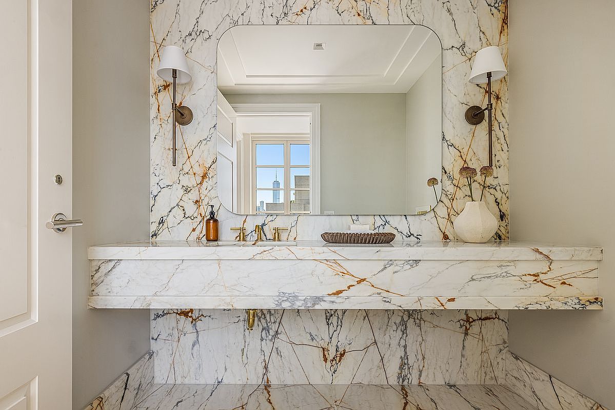 This elegant powder room features a striking vanity clad in dramatic, veined stone that extends from the wall to the floor. A large, rounded rectangular mirror is centered above the vanity, flanked by two classic wall-mounted sconces, while a minimalist vessel sits atop the counter. The space is finished with matching stone surfaces on the wall and floor, creating a cohesive and luxurious aesthetic.
