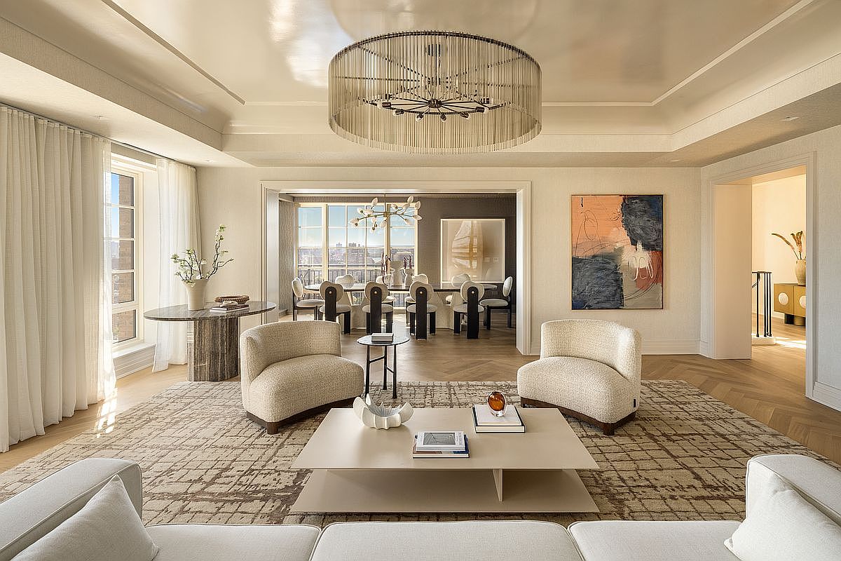 This sophisticated living room features a neutral color palette with textured furnishings, including two curved armchairs and a low-profile coffee table centered on a large patterned rug. A striking tiered chandelier serves as a focal point above the seating area, while a large abstract painting adds a pop of color to the wall. The space flows seamlessly into an adjacent dining room, creating an open and airy atmosphere with herringbone-patterned wood flooring throughout.
