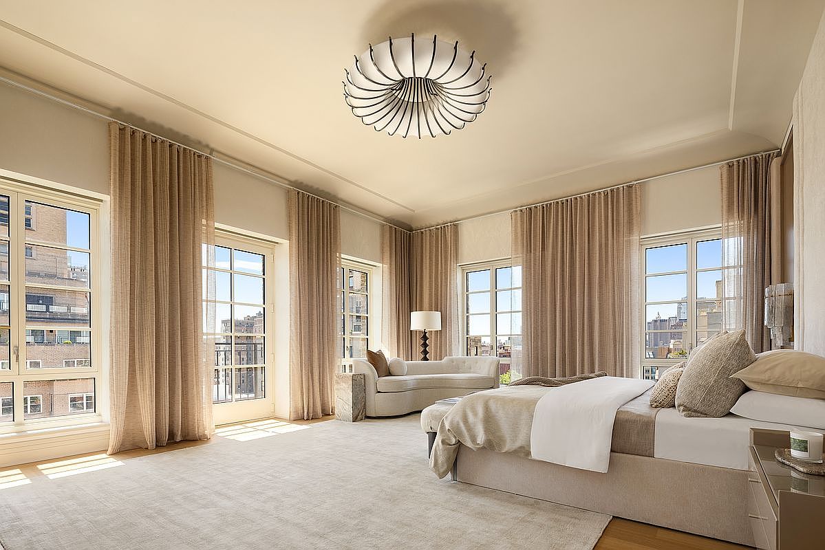 This spacious primary bedroom features a neutral color palette, plush carpeting, and a modern, sculptural chandelier as a central focal point. Large windows with floor-to-ceiling curtains allow for abundant natural light, while a sleek chaise lounge provides a comfortable seating area. The room is elegantly furnished with a king-sized bed and a minimalist nightstand, creating a serene and sophisticated atmosphere.