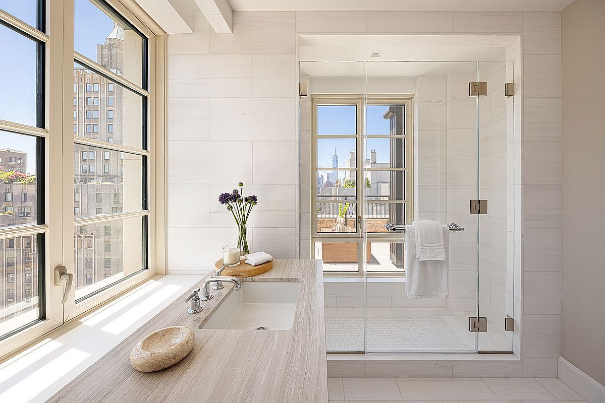 This bright and airy primary bathroom features a sleek vanity with a light-toned stone countertop and an integrated sink, positioned beside a large window that offers an urban view. To the right, a glass-enclosed walk-in shower with light-colored tile walls and a mosaic tile floor creates a seamless, modern aesthetic. The space is filled with natural light, highlighting the clean lines and sophisticated, minimalist design.