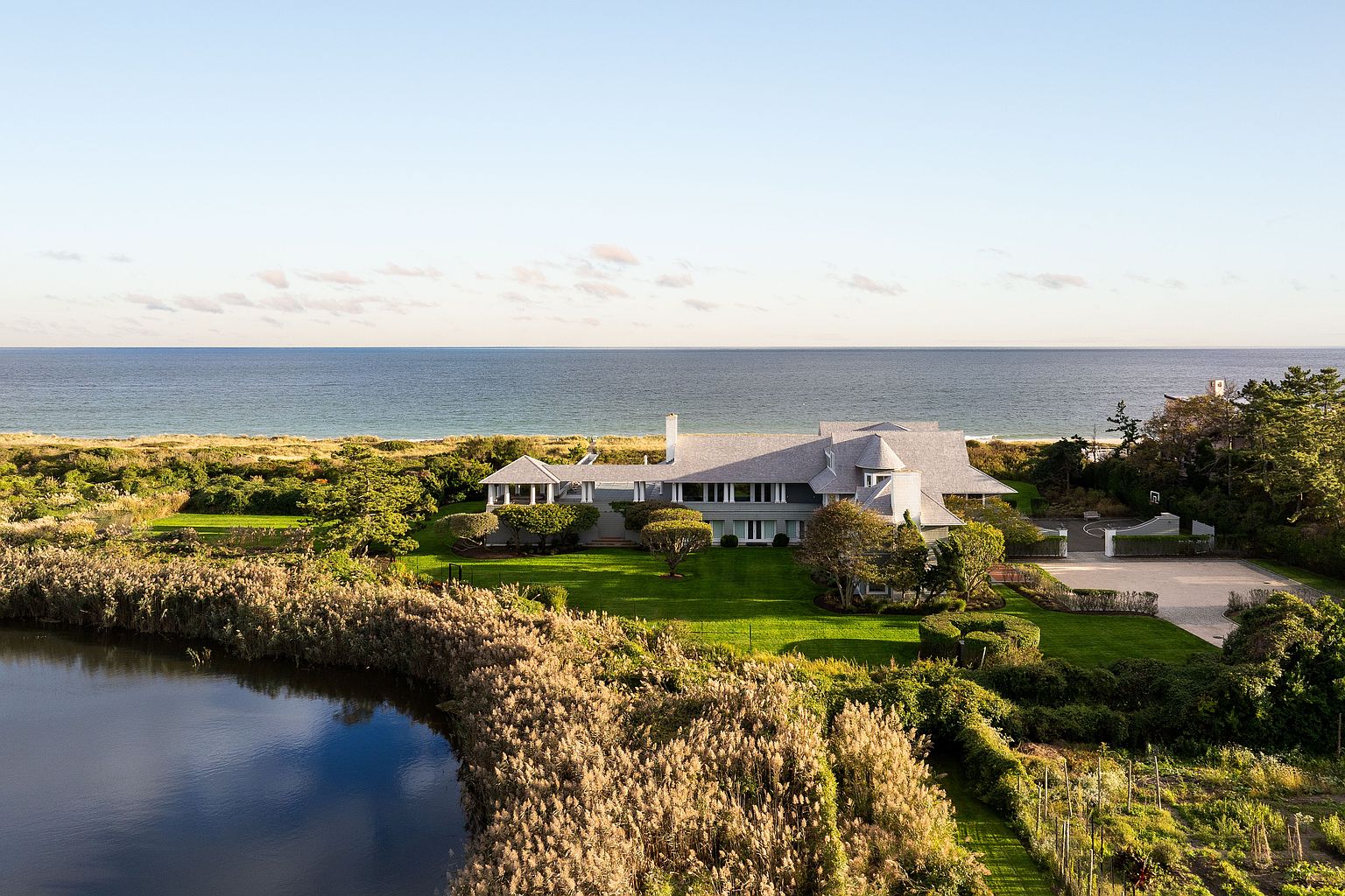 This aerial view showcases a luxurious waterfront estate with meticulously manicured lawns and gardens. The large, gray-shingled house features a complex roofline, a chimney, and multiple windows, suggesting ample natural light. A pond in the foreground adds to the property's serene and private setting, while the ocean provides a stunning backdrop.