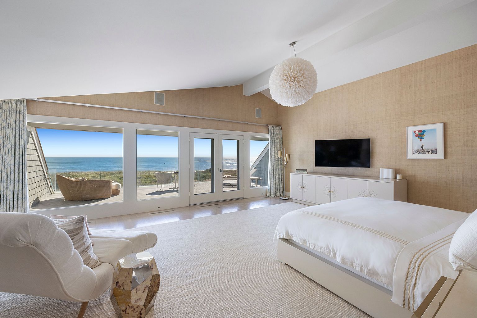 This is a bright and airy primary bedroom with a coastal aesthetic. The room features a large bed with white linens, a seating area with a white chaise lounge, and a wall-mounted television above a white cabinet. Large windows offer stunning ocean views and access to a deck, enhancing the room's serene and luxurious atmosphere.