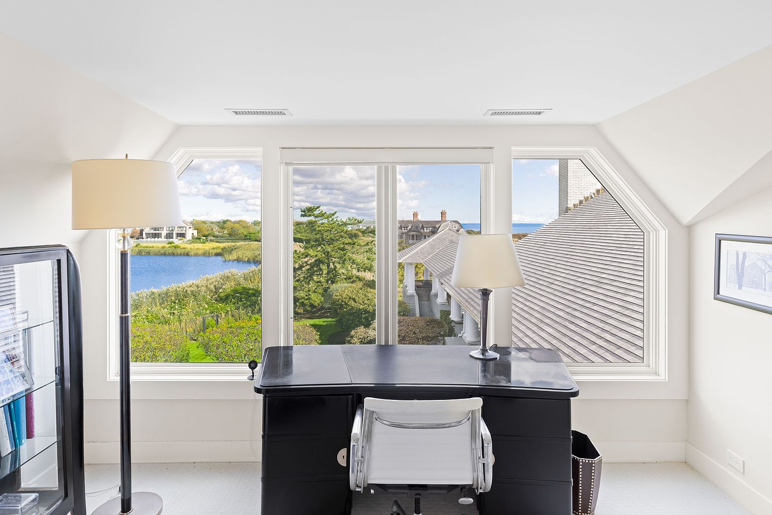 This is an interior shot of a bright and airy office space. The room features a black desk with a white chair, positioned in front of a large window offering a scenic view of a lake and surrounding landscape. A floor lamp stands to the left, and a cabinet is partially visible, contributing to a professional yet comfortable atmosphere.