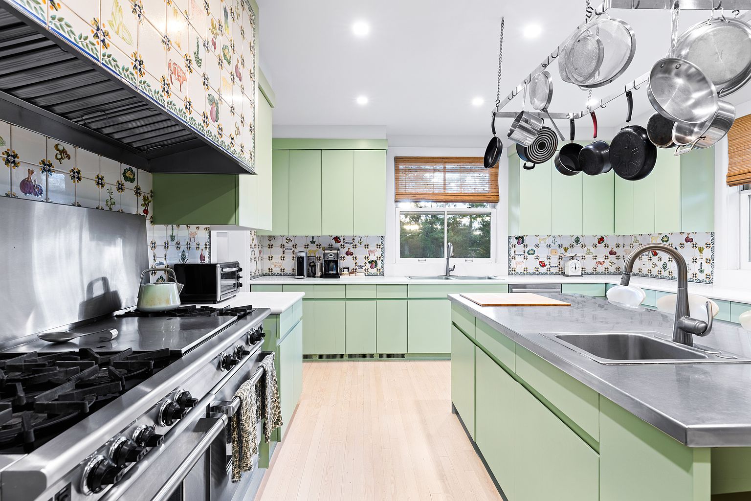 This is a bright and airy kitchen featuring light green cabinetry and a stainless steel island. The backsplash and hood are adorned with decorative tiles, adding a unique touch. Stainless steel appliances and a pot rack above the island complete the functional and stylish space.