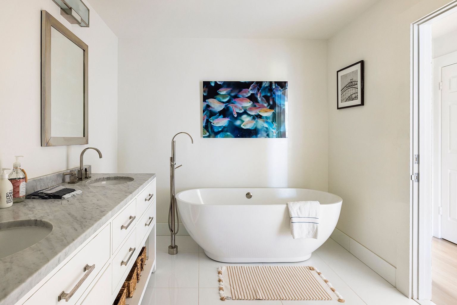 This is a bright and airy primary bathroom featuring a double vanity with a marble countertop and white cabinetry. A freestanding bathtub sits beneath a colorful art piece, complemented by a neutral-toned striped rug. The room is painted in a clean white, enhancing the sense of spaciousness and tranquility.