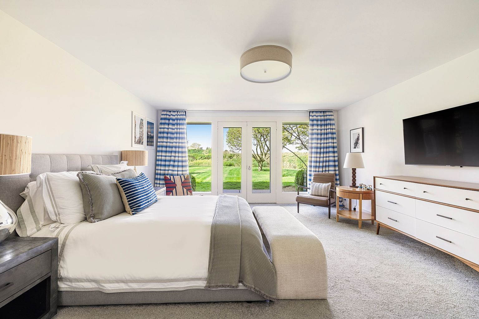 This is a bright and airy primary bedroom featuring a large bed with neutral and blue-striped pillows, a gray upholstered headboard, and a light-colored bench at the foot of the bed. French doors lead to a lush green lawn, framed by blue and white striped curtains. A modern dresser with white drawers and a wooden top sits against the wall, with a large television mounted above it, creating a serene and luxurious atmosphere.