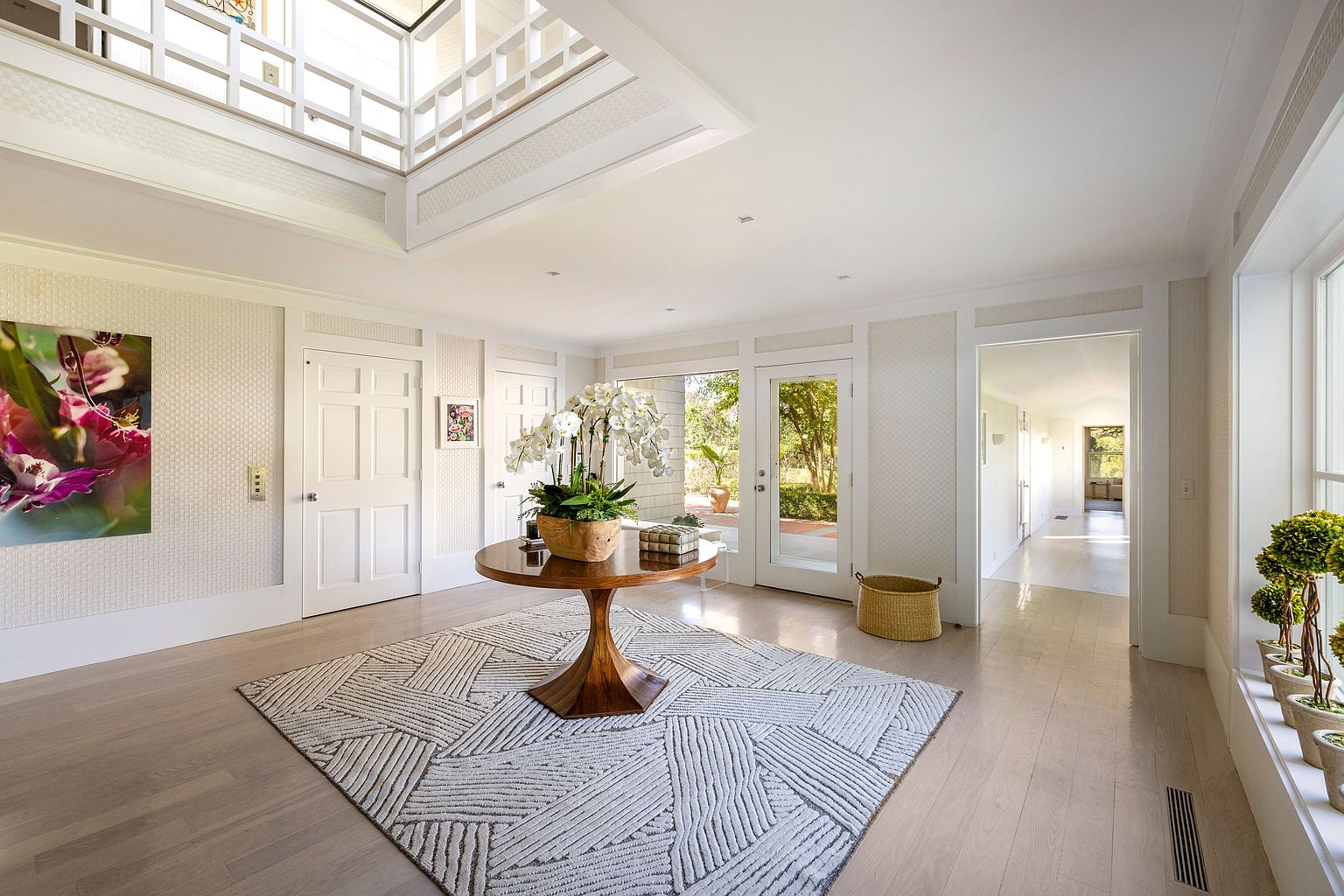 This is a bright and spacious entryway featuring a high ceiling with a skylight and a second-story balcony. The foyer is decorated with a round wooden table topped with a large floral arrangement, set upon a geometric patterned rug. The walls are adorned with a subtle textured wallpaper, and the space is illuminated by natural light streaming through the glass doors leading to the outside.