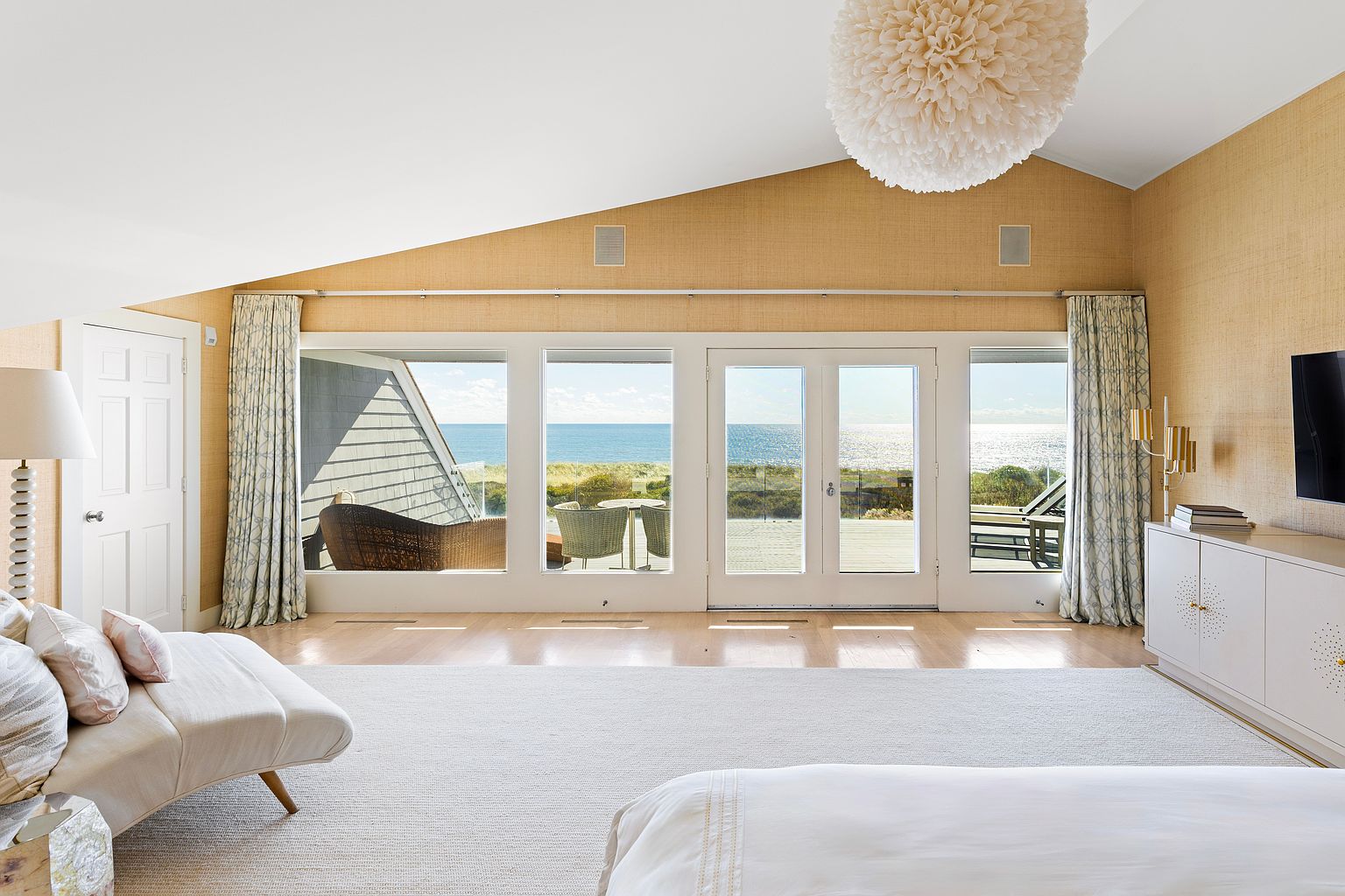 This primary bedroom features a stunning ocean view through large windows and French doors, complemented by light-colored walls and hardwood floors. A plush white rug adds comfort, while a decorative light fixture hangs from the vaulted ceiling. The room is furnished with a chaise lounge and a white dresser, creating a serene and luxurious atmosphere.