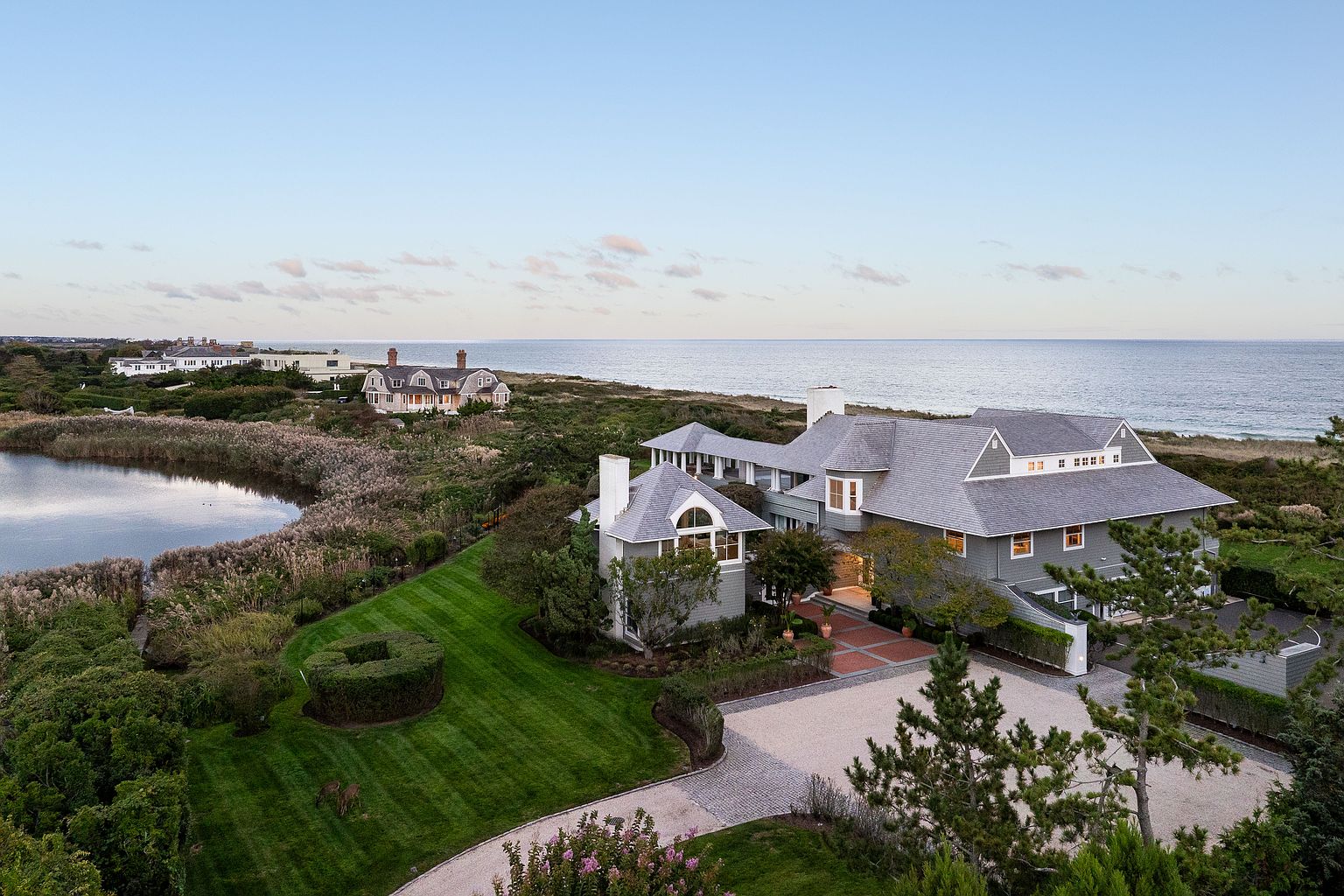 This aerial view showcases a luxurious coastal estate with meticulously manicured lawns and mature landscaping. The large, gray-shingled home features multiple gables, a brick driveway, and a serene pond nearby. The property offers stunning ocean views, emphasizing its prime location and high-end appeal.
