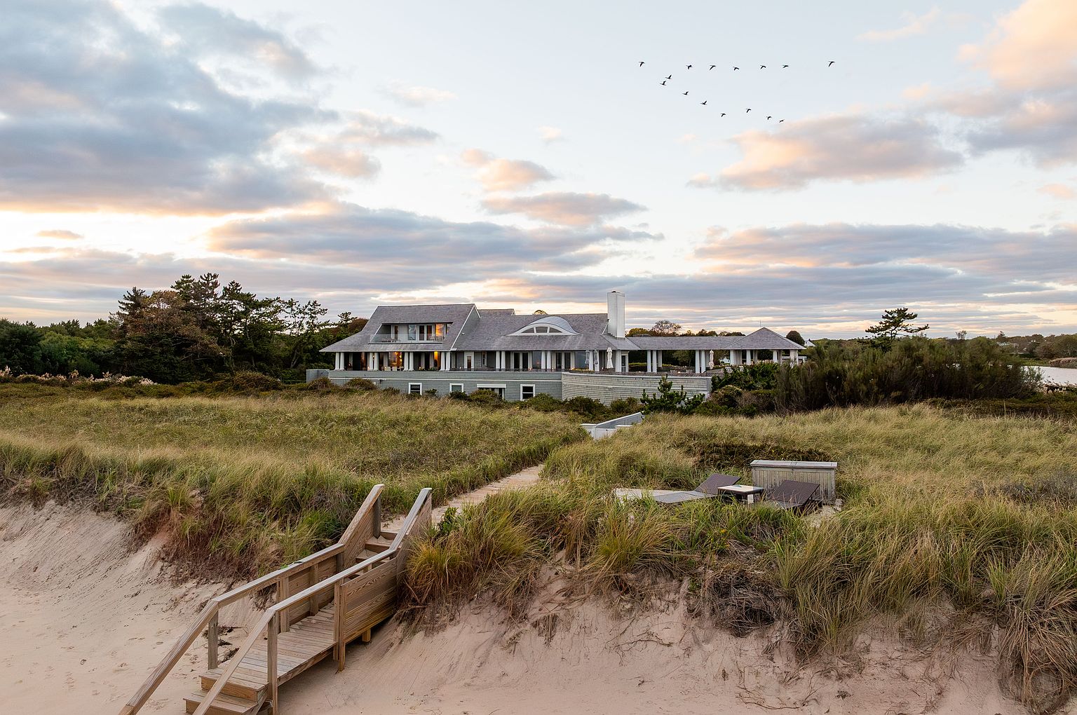 This image showcases the rear exterior of a large, luxurious home situated near a beach. The house features a gray roof, light green siding, and multiple windows, with a sprawling lawn leading down to a sandy beach accessible via wooden stairs. The scene is set during a tranquil evening, with a flock of birds flying overhead, creating a serene and upscale coastal living impression.