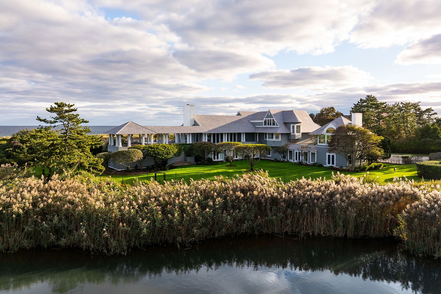 This aerial view showcases a sprawling, luxurious home with a gray roof and light blue siding, nestled amidst lush greenery and overlooking a body of water. The property features a well-manicured lawn, mature trees, and tall reeds along the water's edge, creating a serene and private setting. The architectural style is elegant and timeless, suggesting a high-end coastal estate.
