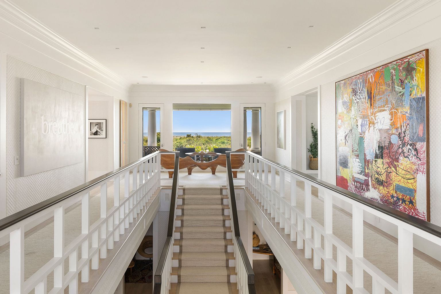 This interior shot showcases a bright and airy hallway with a staircase leading down. The hallway features white walls, white railings with dark trim, and artwork on the walls, creating a sophisticated and inviting atmosphere. The view through the windows at the end of the hallway reveals a scenic coastal landscape, enhancing the property's appeal.