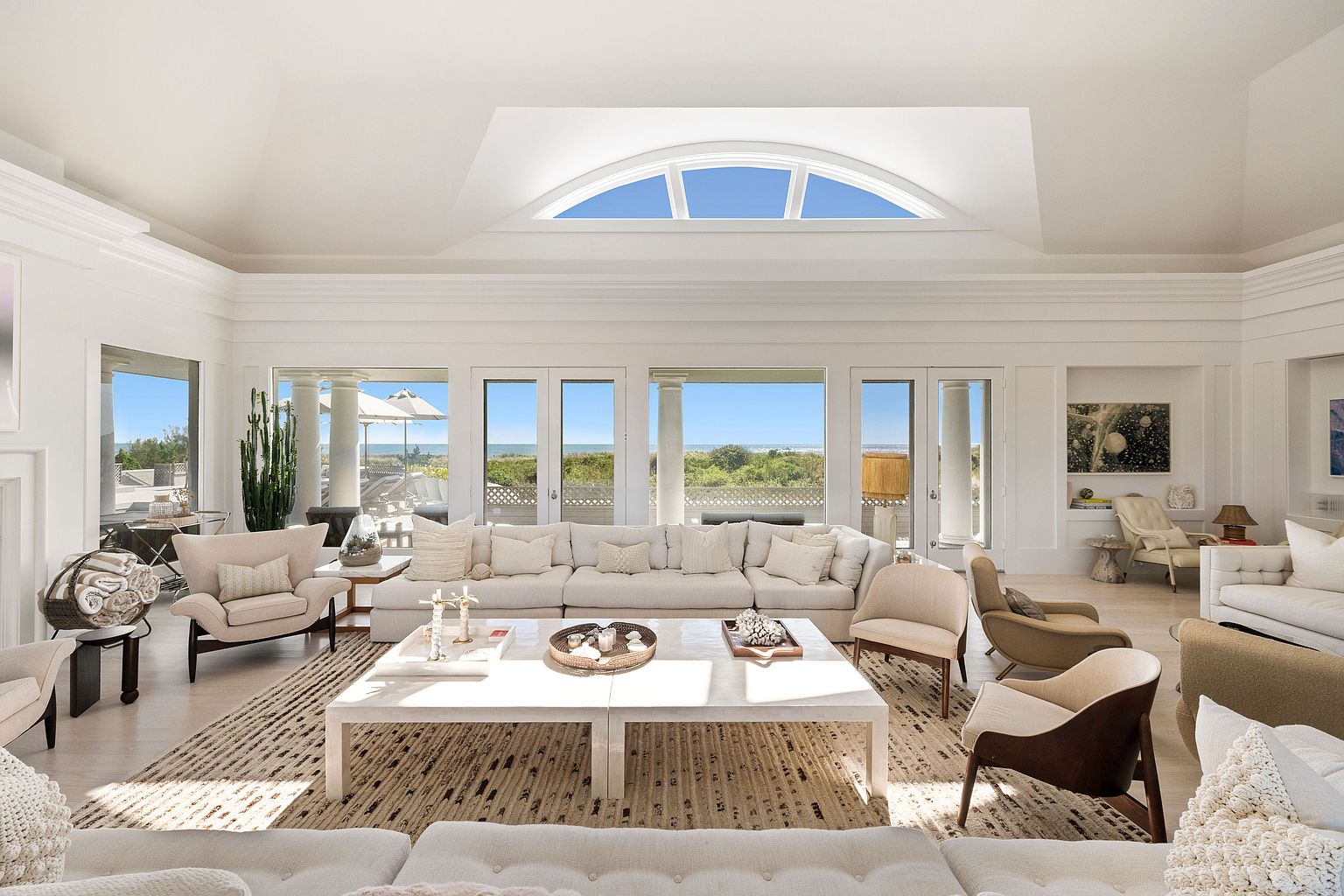 This is a bright and spacious living room featuring a large white sectional sofa, a light-colored area rug, and a white coffee table. The room has large windows and a skylight that offer views of the outdoors and flood the space with natural light. The overall style is elegant and coastal, creating a serene and inviting atmosphere.