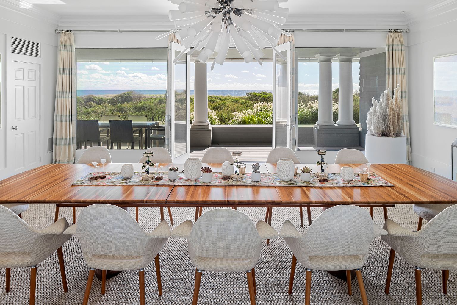 This is an interior shot of a dining room featuring a long wooden table with white chairs. The table is decorated with a floral runner and various decorative items. Large glass doors open to a patio with a view of the ocean, creating a bright and airy atmosphere. The room is well-lit with a modern chandelier, and the overall style is elegant and coastal.