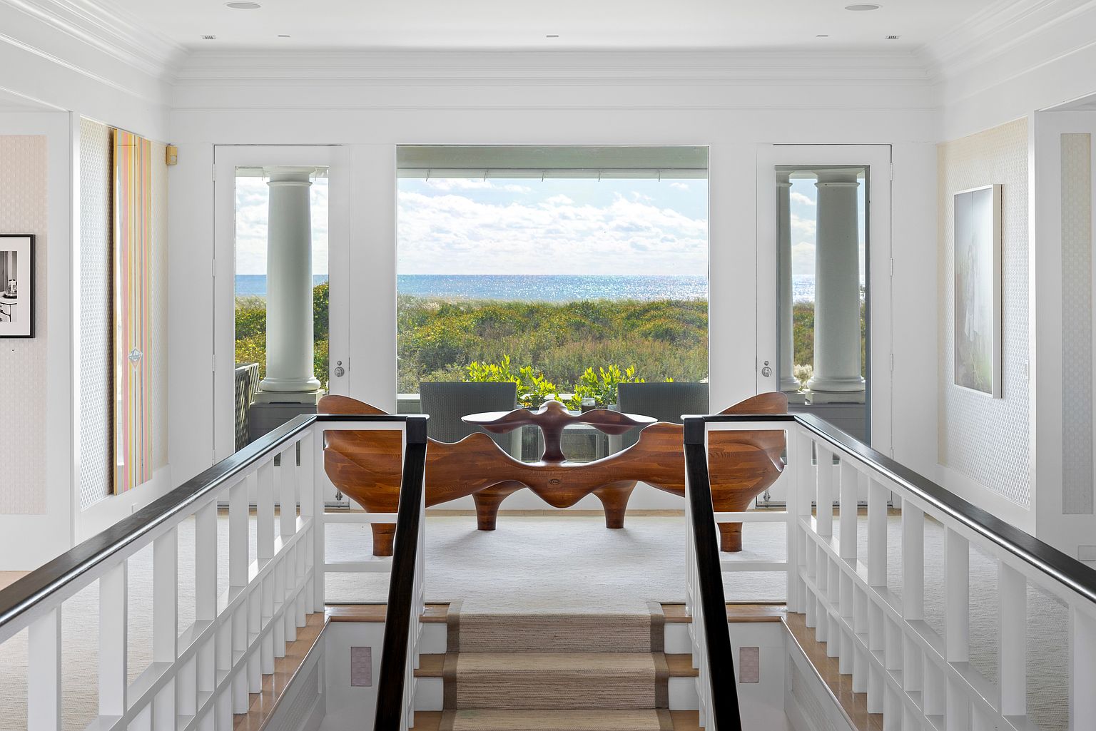 This interior shot showcases a bright hallway with stairs leading down, offering a glimpse of the ocean through a large window. The hallway features white walls, white railings with black accents, and a unique wooden table or bench in the distance, creating a sophisticated and inviting atmosphere. The view of the ocean adds a luxurious touch, emphasizing the property's prime location.