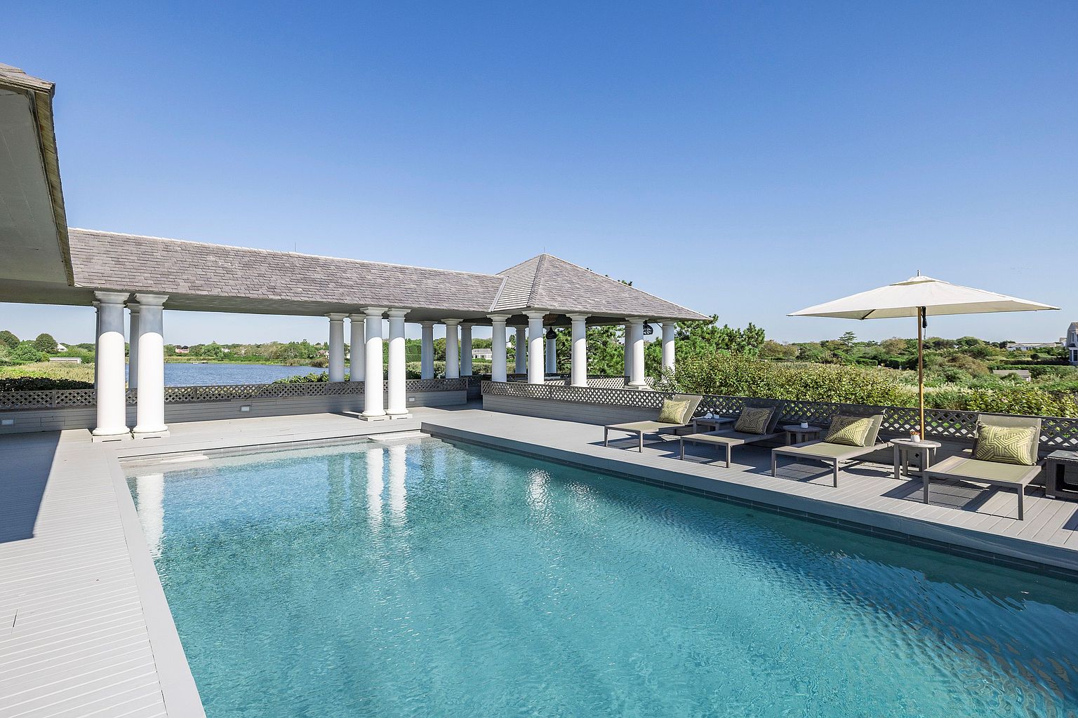 This image showcases a luxurious outdoor pool area with a covered pavilion featuring white columns and a gray roof. The pool's clear blue water reflects the sky, and lounge chairs with cushions are arranged on the deck alongside a white umbrella. The surrounding landscape includes lush greenery and a glimpse of a body of water, creating a serene and upscale atmosphere.