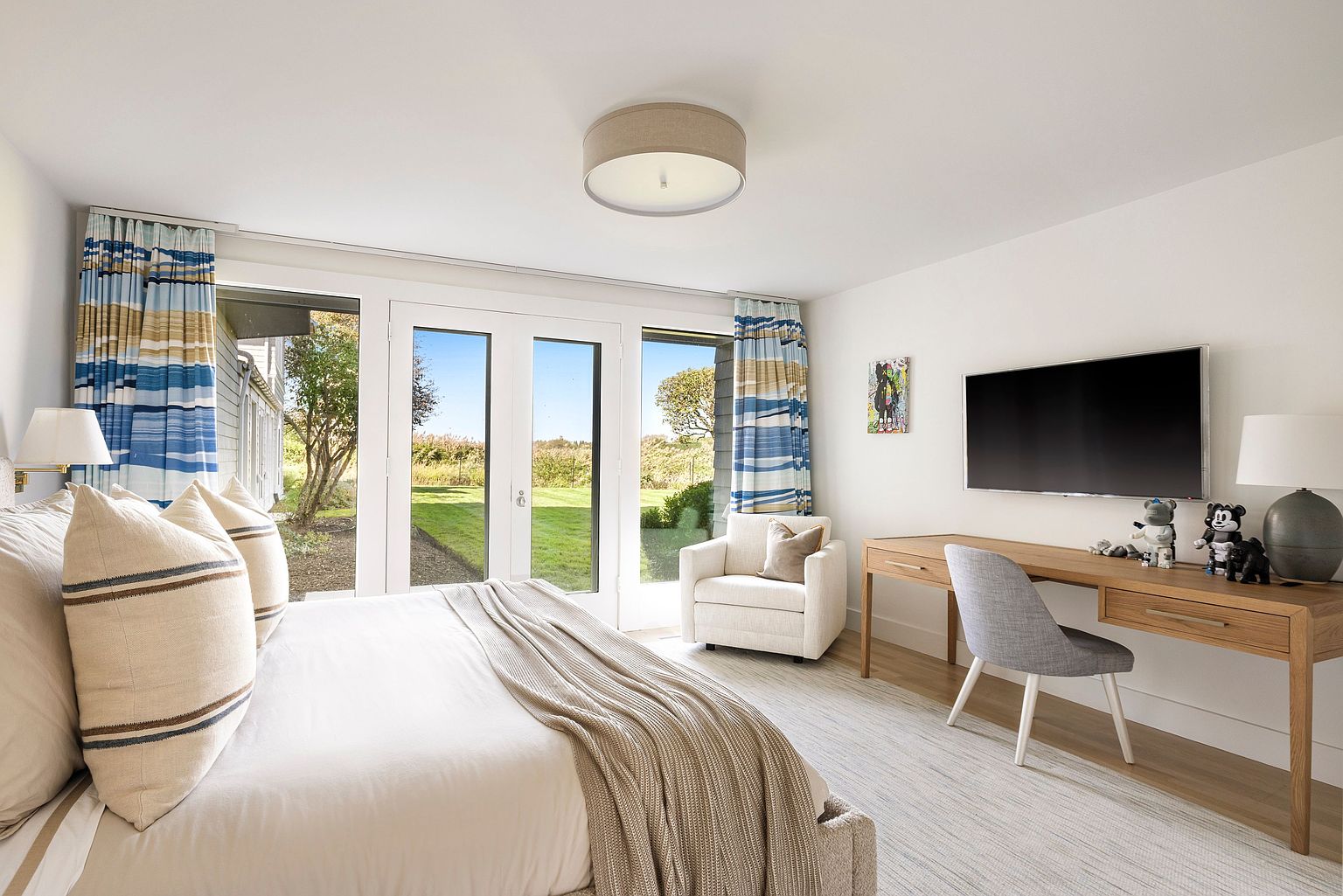 This is a bright and airy primary bedroom featuring a king-size bed with neutral-toned linens and decorative pillows. French doors lead to an outdoor space, framed by stylish curtains. A desk area with a modern chair and wall-mounted television provides a functional workspace, enhancing the room's appeal.