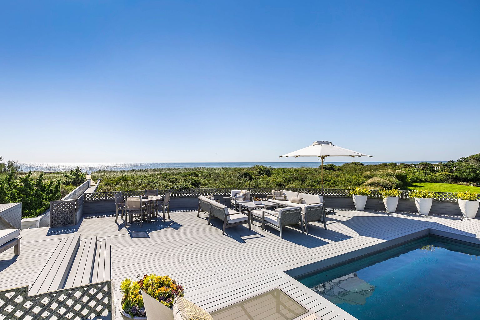 This image showcases a luxurious outdoor deck area with a stunning ocean view. The deck features comfortable lounge seating, a dining table, and an umbrella for shade, all arranged on a gray-toned wooden surface. A swimming pool is adjacent to the deck, and the surrounding landscape includes lush greenery and dunes, creating a serene and private atmosphere.