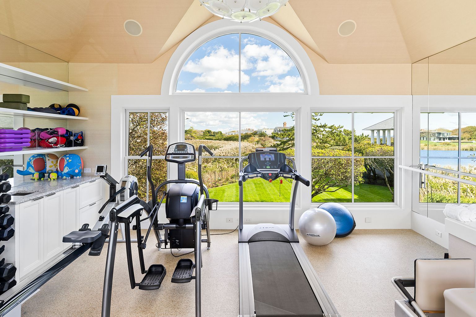 This is a well-lit home gym featuring a treadmill, elliptical machine, and exercise balls. Large windows offer a view of the outdoors, bringing natural light into the space. The room includes built-in storage and a neutral color palette, creating a clean and inviting workout environment.