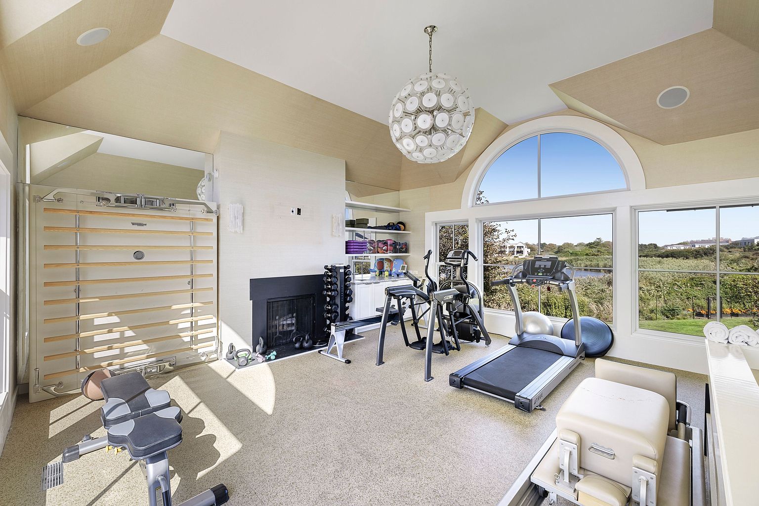This is a well-equipped home gym featuring a variety of exercise machines, including a treadmill, elliptical, rowing machine, and weight bench. The room has large windows offering natural light and views of the outdoors, complemented by a modern chandelier. The space also includes a wall-mounted ladder and a fireplace, creating a unique and inviting workout environment.