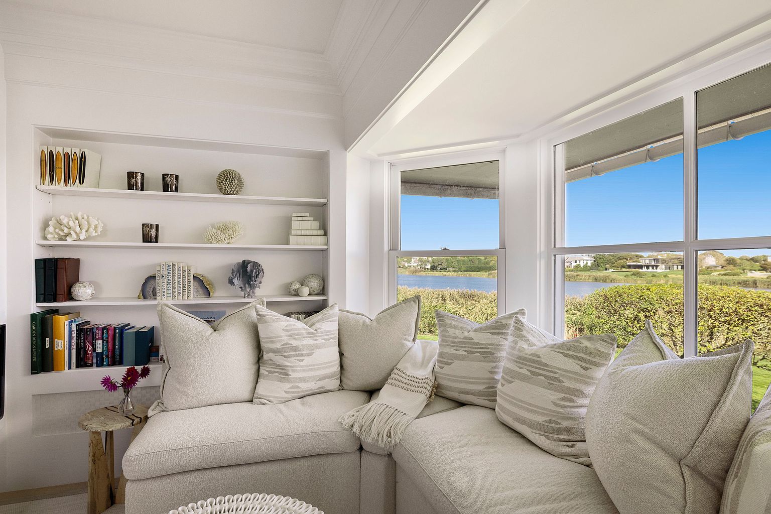 This inviting living room features a built-in bookshelf filled with decorative items and books, adding character to the space. A comfortable, neutral-toned sectional sofa is adorned with patterned pillows and a throw blanket, creating a cozy atmosphere. Large windows offer a serene view of the water and landscape, enhancing the room's appeal.