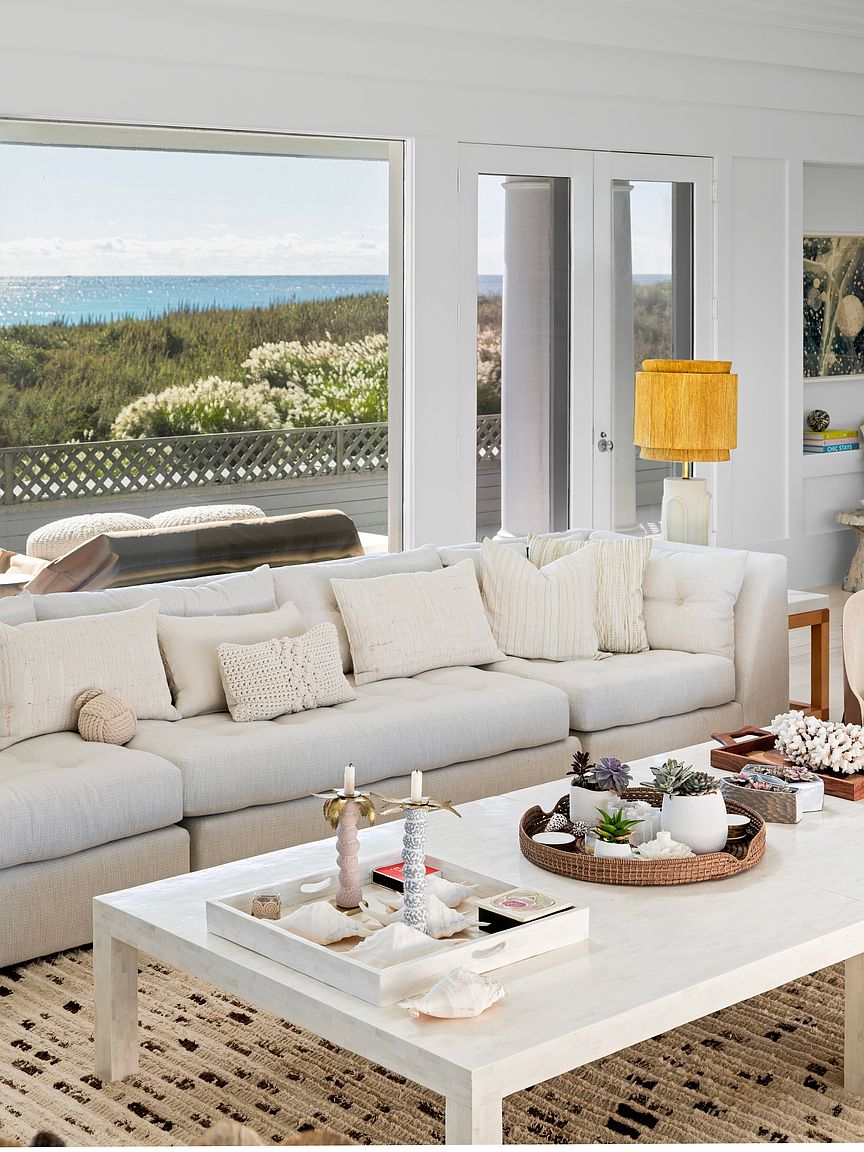 This is an inviting living room featuring a large, comfortable sectional sofa adorned with neutral-toned pillows. A white coffee table displays decorative trays and objects, while a textured rug adds warmth to the space. Large windows offer a view of the ocean and natural light, enhancing the room's serene and coastal aesthetic.