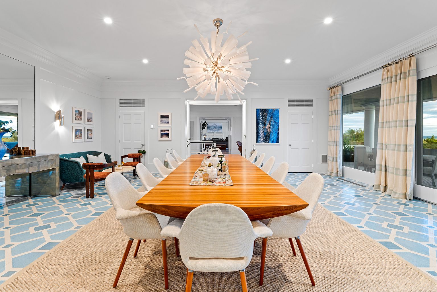 This is a bright and spacious dining room featuring a long wooden table surrounded by modern white chairs. A unique, sculptural chandelier hangs above the table, providing ample lighting. The room is decorated in a clean, contemporary style with white walls and a patterned blue and white floor, creating an elegant and inviting atmosphere.