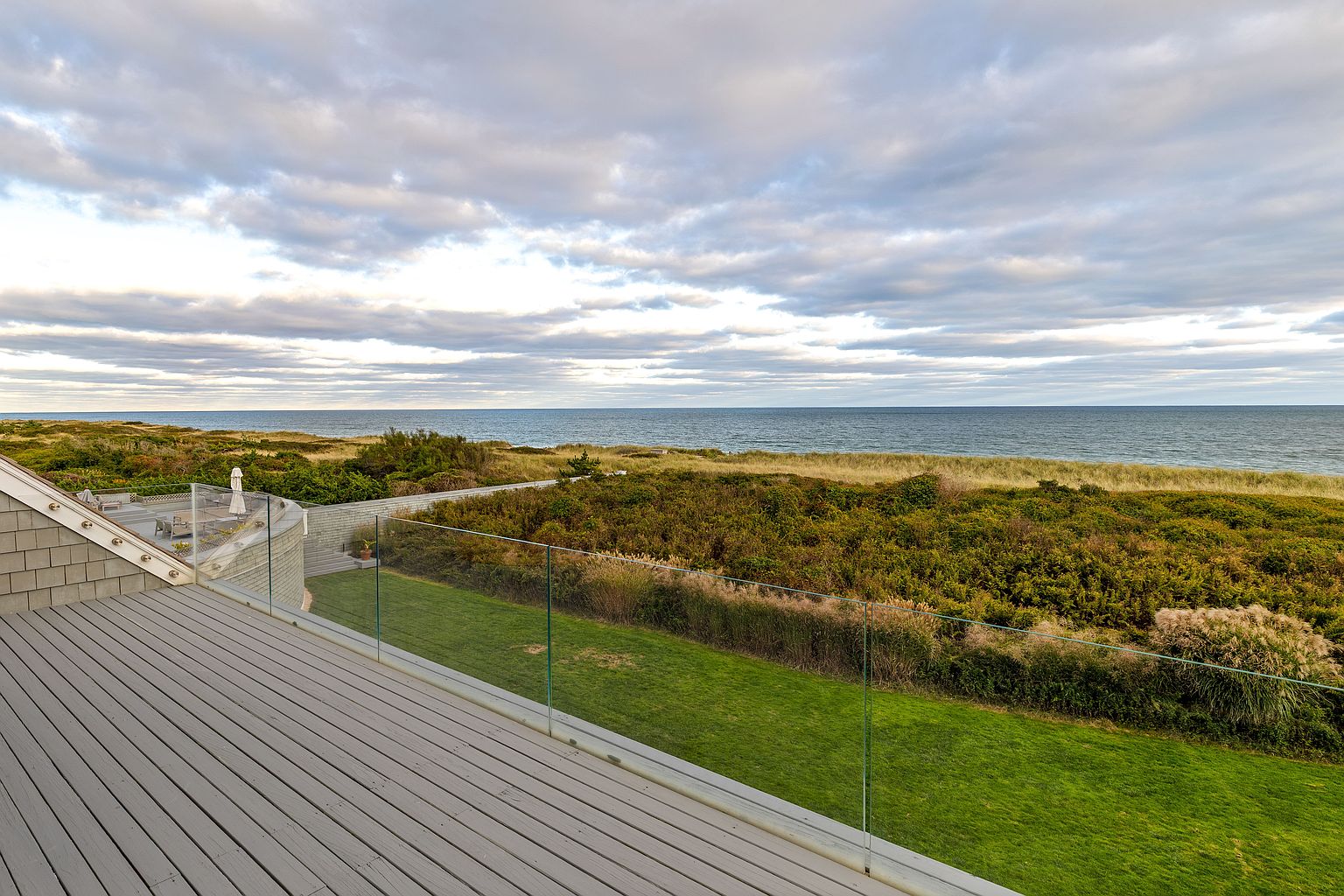 This image showcases a spacious outdoor deck or patio area with a modern glass railing, offering an unobstructed view of the ocean and surrounding dunes. The deck is constructed with gray-toned wood, and the landscape features lush green grass and natural vegetation. The scene is set under a cloudy sky, creating a serene and inviting atmosphere.