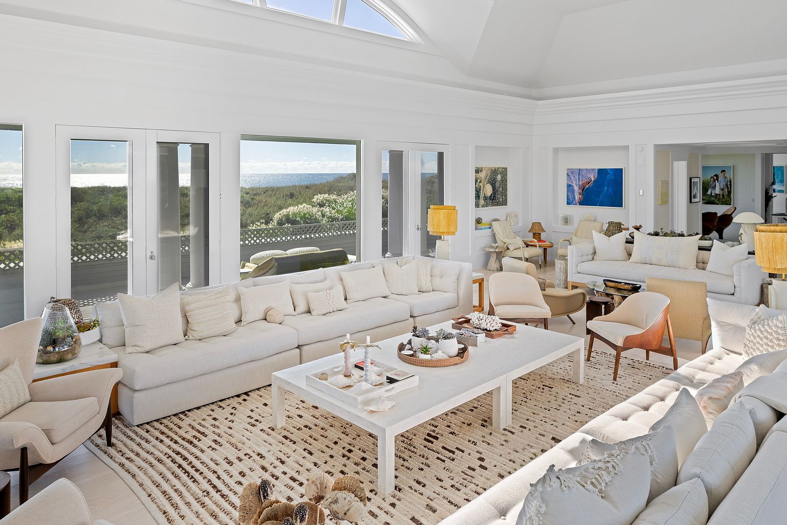 This is a bright and airy living room featuring a large white sectional sofa adorned with numerous pillows, a light-colored area rug with a subtle pattern, and a white coffee table styled with decorative objects. Large windows offer views of the ocean and surrounding greenery, enhancing the room's serene and luxurious atmosphere. The room is spacious and well-lit, creating an inviting and comfortable living space.