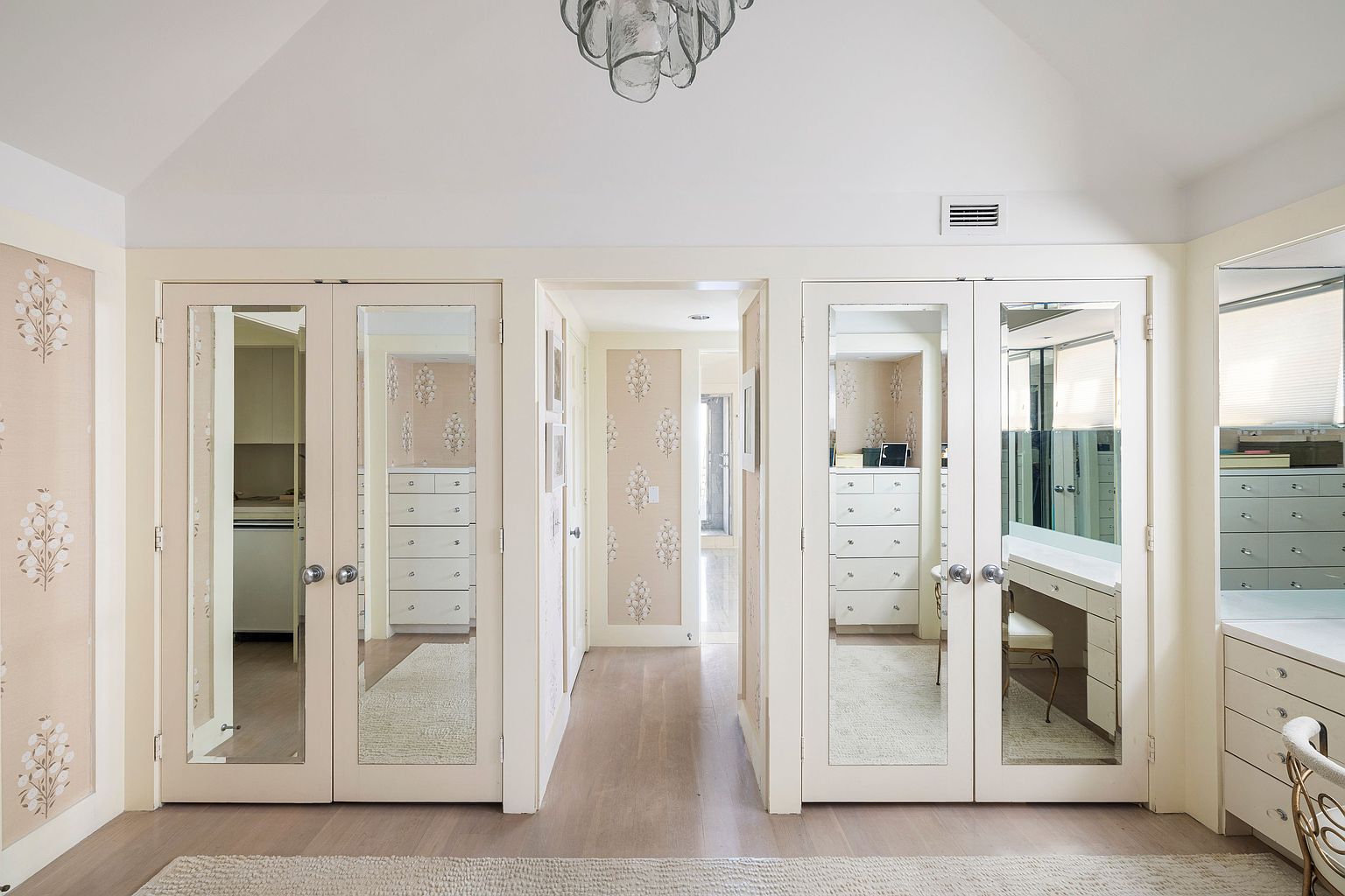 This is an interior shot of a luxurious walk-in closet. The closet features mirrored doors, custom cabinetry, and elegant wallpaper. The space is well-lit and organized, creating a sophisticated and functional dressing area. The perspective is from the entrance of the closet, showcasing the depth and layout of the space.