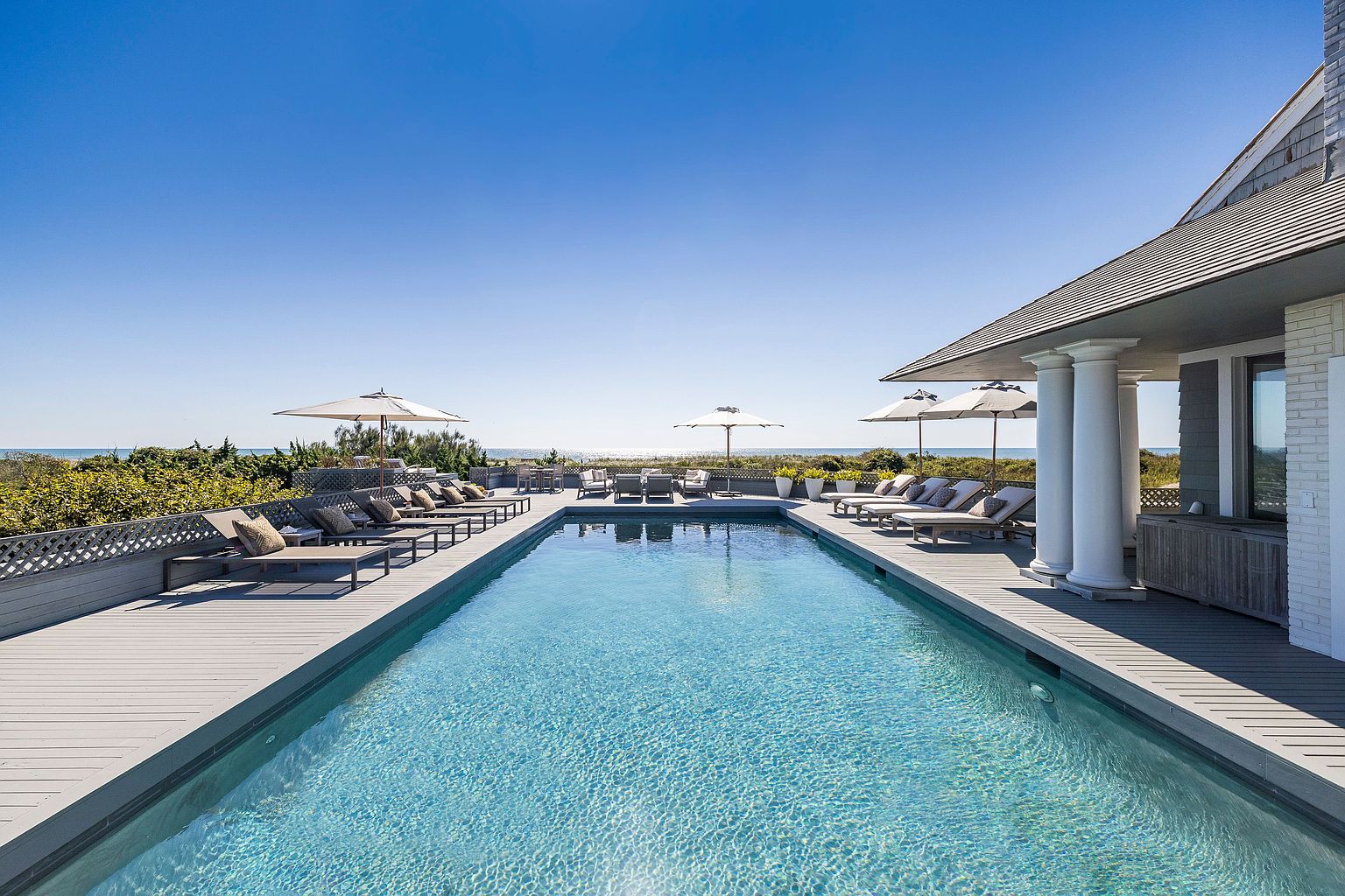 This image showcases a luxurious outdoor pool area, featuring a long, rectangular pool with clear blue water. The pool is surrounded by a wooden deck furnished with lounge chairs and umbrellas, creating a relaxing space. The property overlooks a beach and ocean, offering stunning views and enhancing the appeal of this high-end real estate.