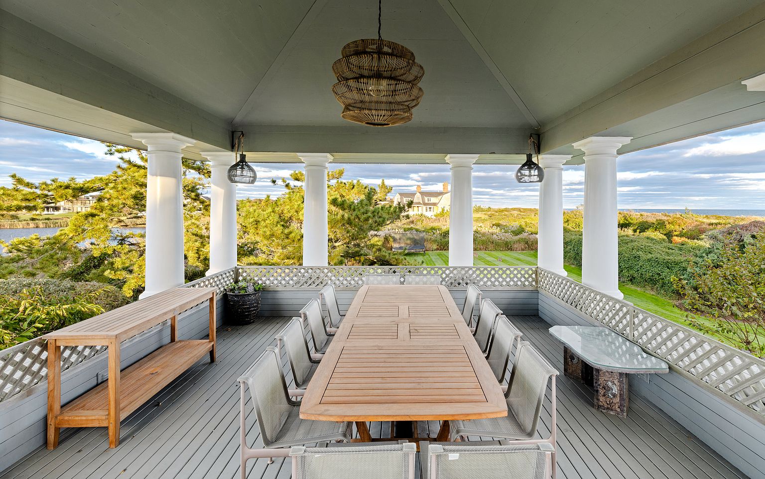 This is a spacious outdoor dining area on a covered patio or deck. The focal point is a large wooden dining table surrounded by chairs, suggesting ample seating for gatherings. The area is framed by white columns and a decorative railing, offering views of the surrounding landscape and creating an inviting atmosphere for outdoor entertaining.