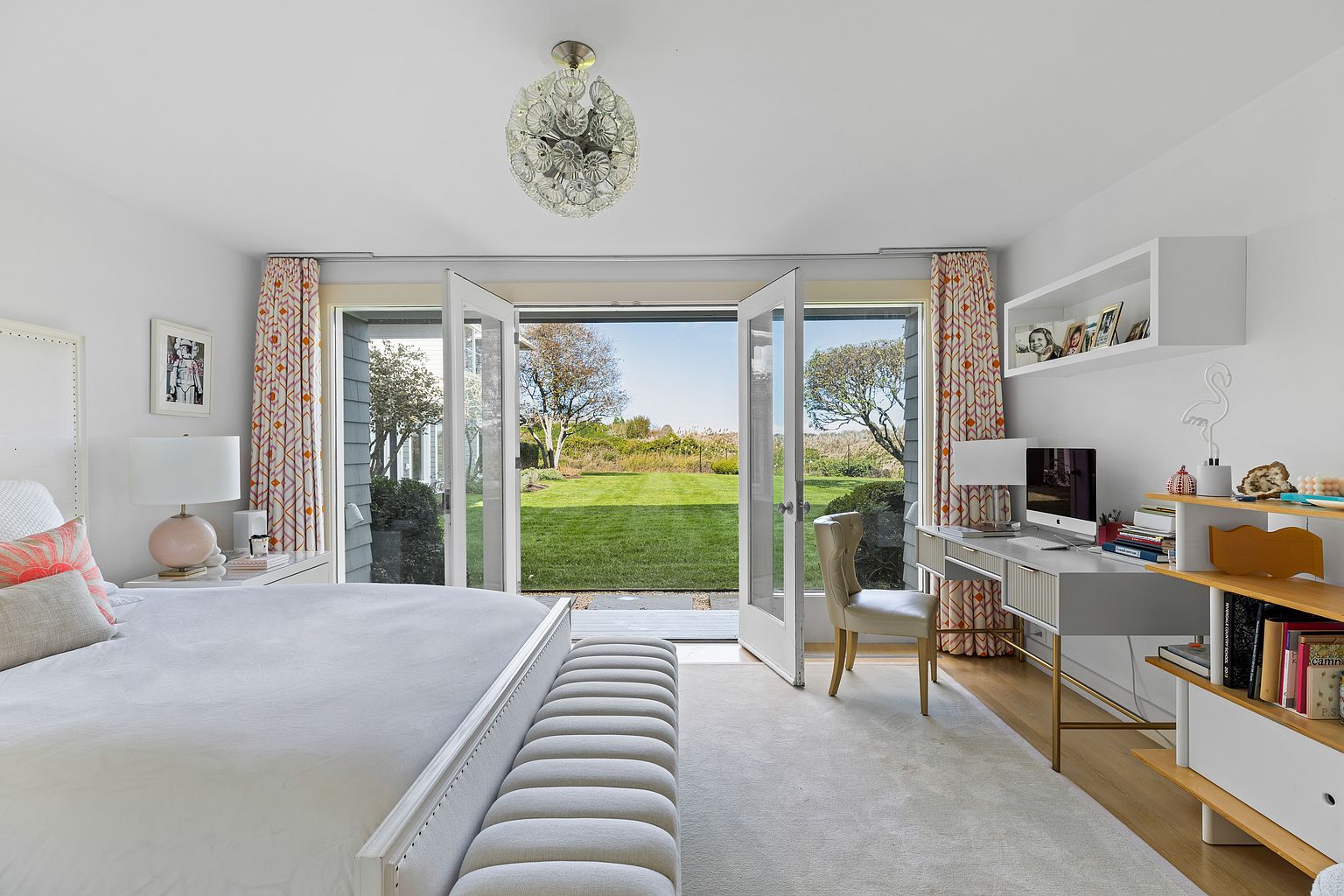 This is a bright and airy primary bedroom featuring a large bed with a white frame and a tufted bench at the foot. French doors open to a lush green lawn, bringing the outdoors in. A desk area with a computer and shelving provides a functional workspace, enhancing the room's appeal.