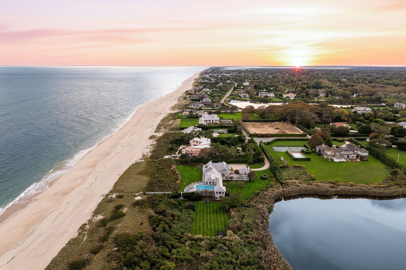This aerial view showcases a stunning coastal property featuring a large, modern home with a pool overlooking the ocean. The manicured lawn extends to a private beach, while a serene pond adds to the landscape's charm. The overall impression is one of luxury and exclusivity, perfect for a high-end real estate listing.