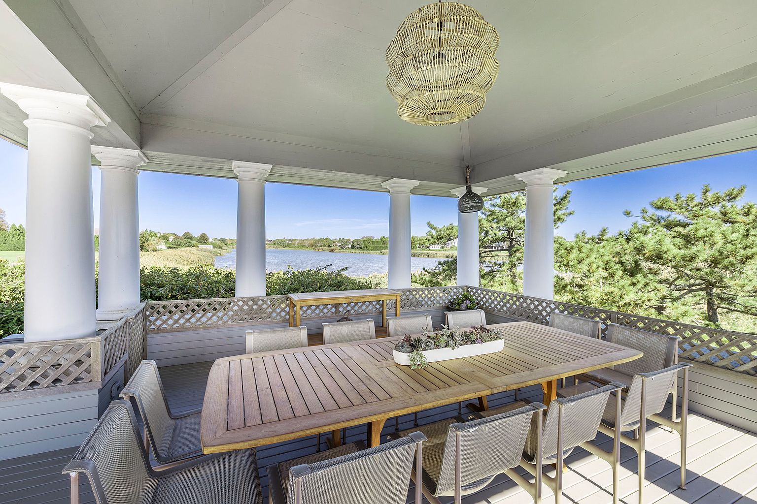 This image showcases a beautifully designed outdoor patio or deck area, featuring a large wooden dining table with seating for ten, perfect for entertaining. The space is covered and supported by elegant white columns, offering shade and a sense of grandeur, while the lattice railing provides a charming detail. The view overlooks a serene body of water, enhancing the appeal of this outdoor living space.
