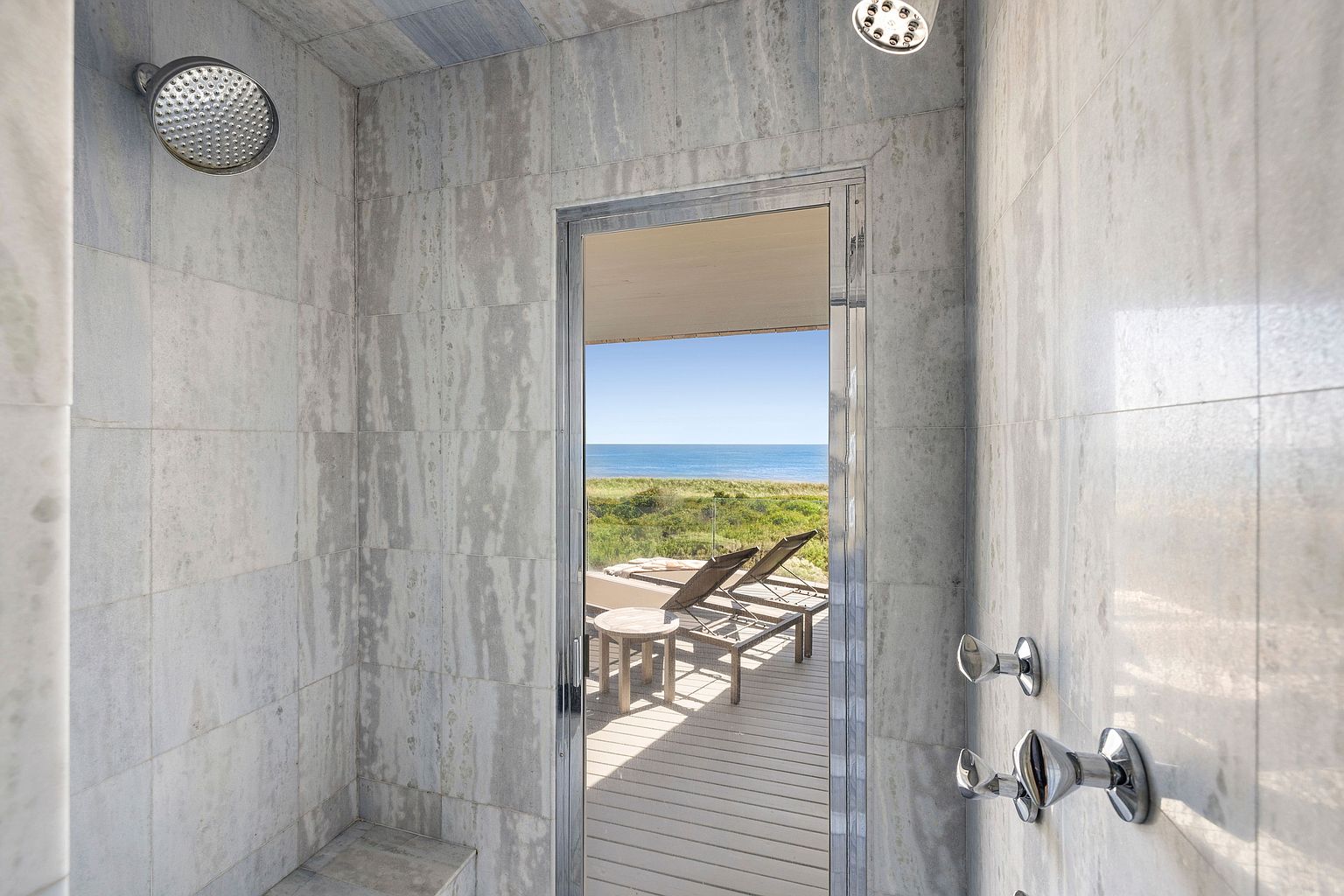 This is a luxurious primary bathroom featuring a walk-in shower with marble-like tiling and modern fixtures. The shower opens to an outdoor deck with lounge chairs and an ocean view, creating a seamless indoor-outdoor experience. The perspective is from inside the shower, looking out towards the deck and ocean.