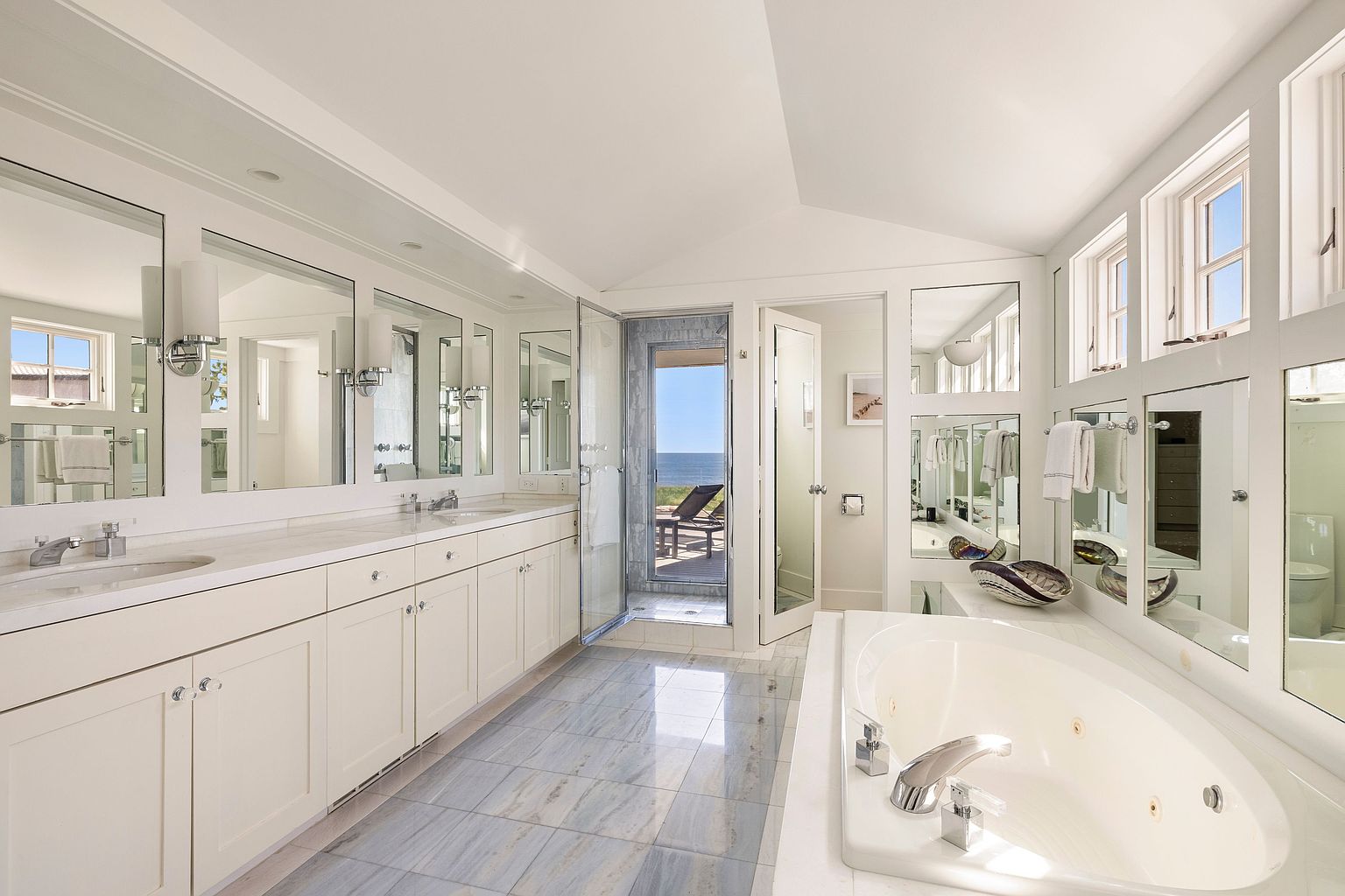 This is a luxurious primary bathroom featuring a long vanity with multiple sinks and mirrors, a large soaking tub, and a glass-enclosed shower with a view of the ocean. The bathroom has a bright and airy feel, with white cabinetry and light-colored tile flooring. The design emphasizes relaxation and tranquility.