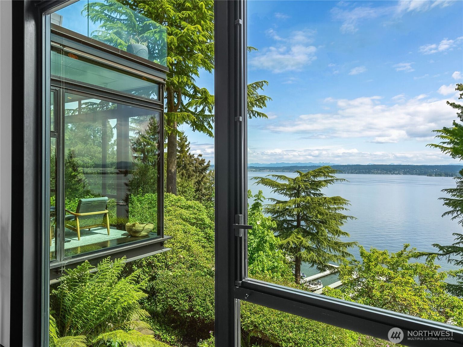 This view captures a modern architectural space featuring expansive, floor-to-ceiling glass walls that frame a lush, tree-filled landscape and a serene waterfront. A single chair sits within a glass-enclosed nook, creating a tranquil reading or observation area that blurs the line between indoor living and the natural environment. The dark window frames provide a sharp, contemporary contrast to the vibrant greenery and the expansive blue sky visible outside.
