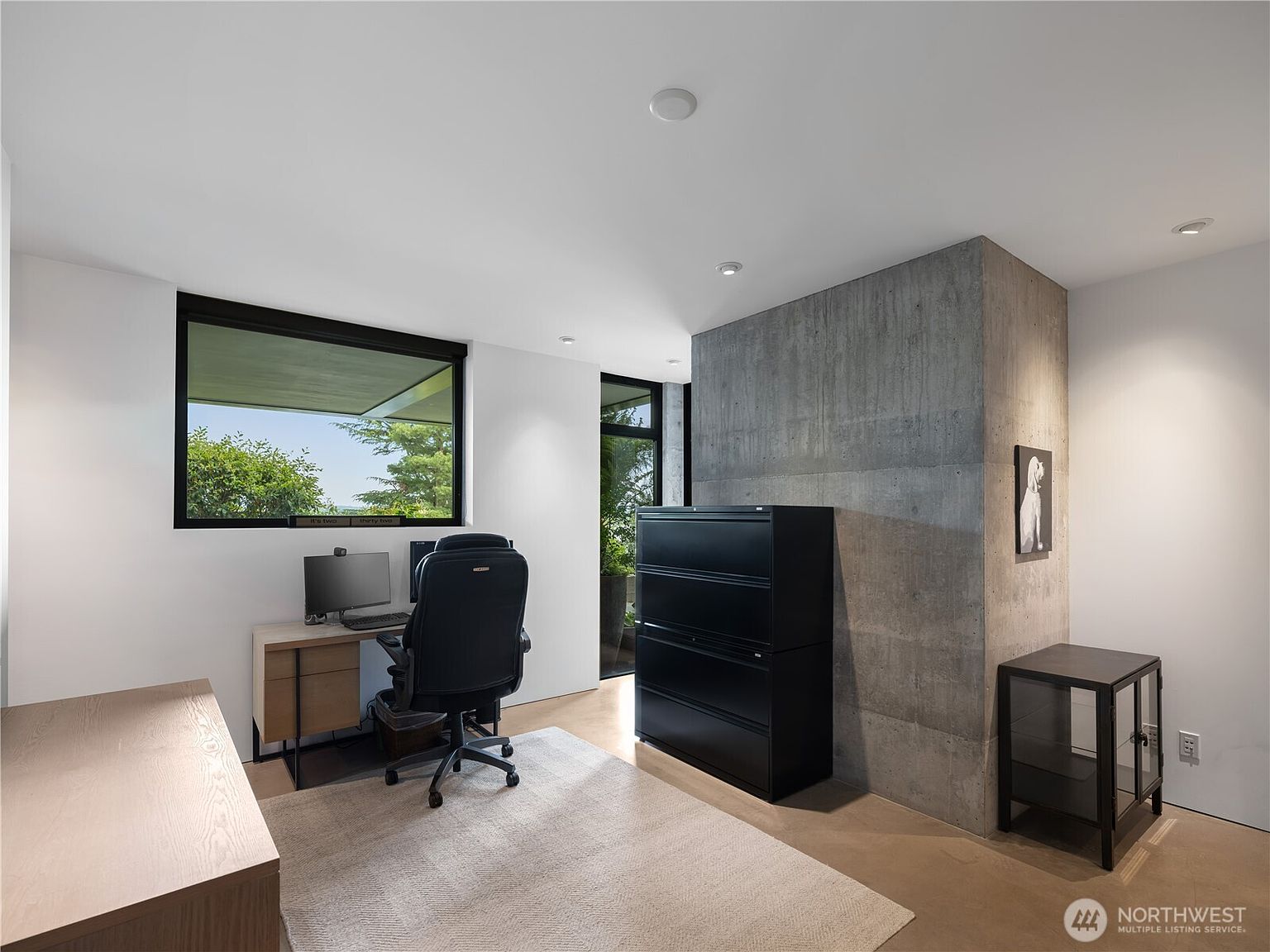 This modern home office features a minimalist aesthetic with a prominent raw concrete wall serving as a focal point. The room is furnished with a light wood desk, a black ergonomic office chair, and a tall black filing cabinet, all set atop a neutral area rug. A large window provides natural light and a view of the outdoors, while recessed lighting illuminates the clean, white-walled space.