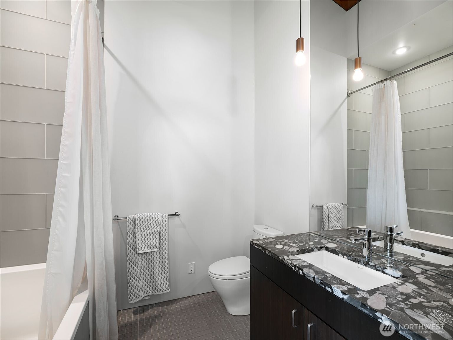This modern bathroom features a striking vanity with a dark, patterned stone countertop and a rectangular undermount sink. The space includes a toilet, a shower area with a white curtain, and a large mirror reflecting the room, all accented by two hanging pendant lights. The walls are finished in a neutral tone, complemented by light-colored rectangular wall tiles in the shower area and small square floor tiles.