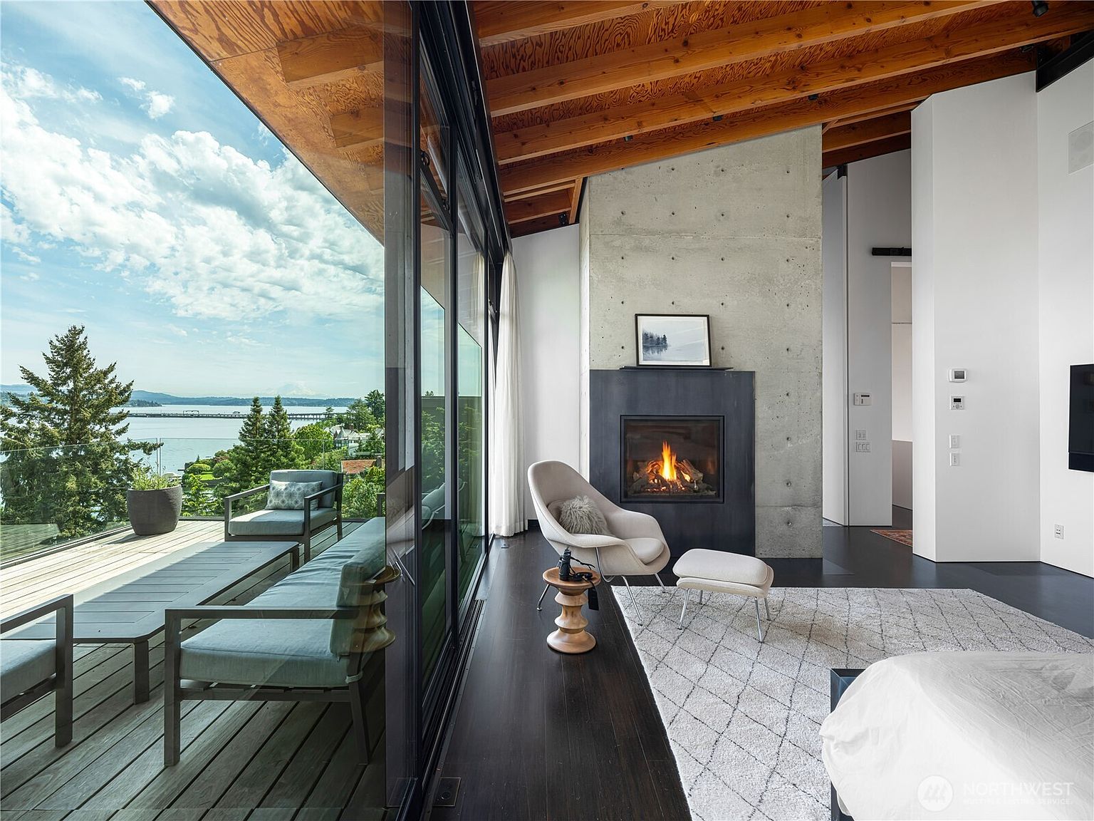 This sophisticated primary bedroom features a striking concrete chimney breast housing a modern fireplace, set against a backdrop of exposed wood ceiling beams. A wall of floor-to-ceiling glass sliding doors opens onto a spacious deck, offering panoramic views of the water and surrounding trees. The room is furnished with a contemporary lounge chair, a small wooden side table, and a plush area rug, creating a serene and luxurious atmosphere.