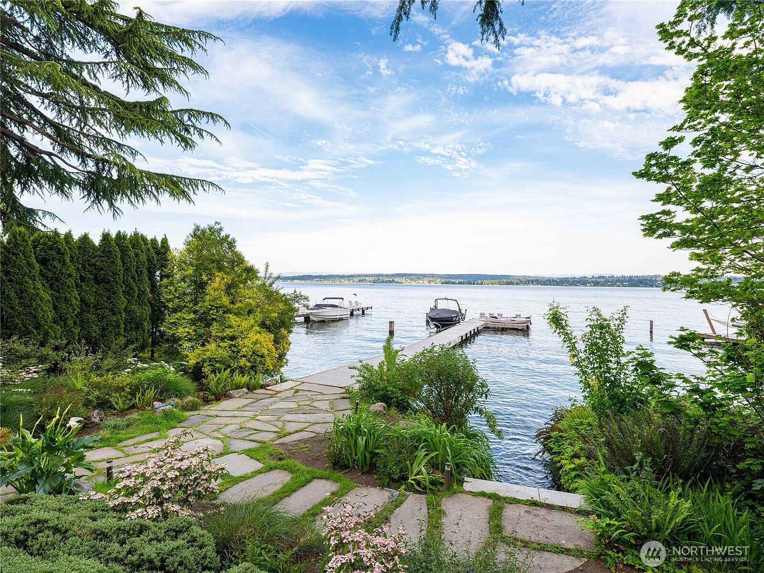 This scenic waterfront property features a stone-paved pathway leading through a lush, landscaped garden toward a private wooden dock extending into a calm lake. The foreground is filled with vibrant greenery and flowering shrubs, while the background showcases a wide, open view of the water under a bright, partly cloudy sky. Two boats are moored at the dock, emphasizing the property's direct access to recreational water activities.